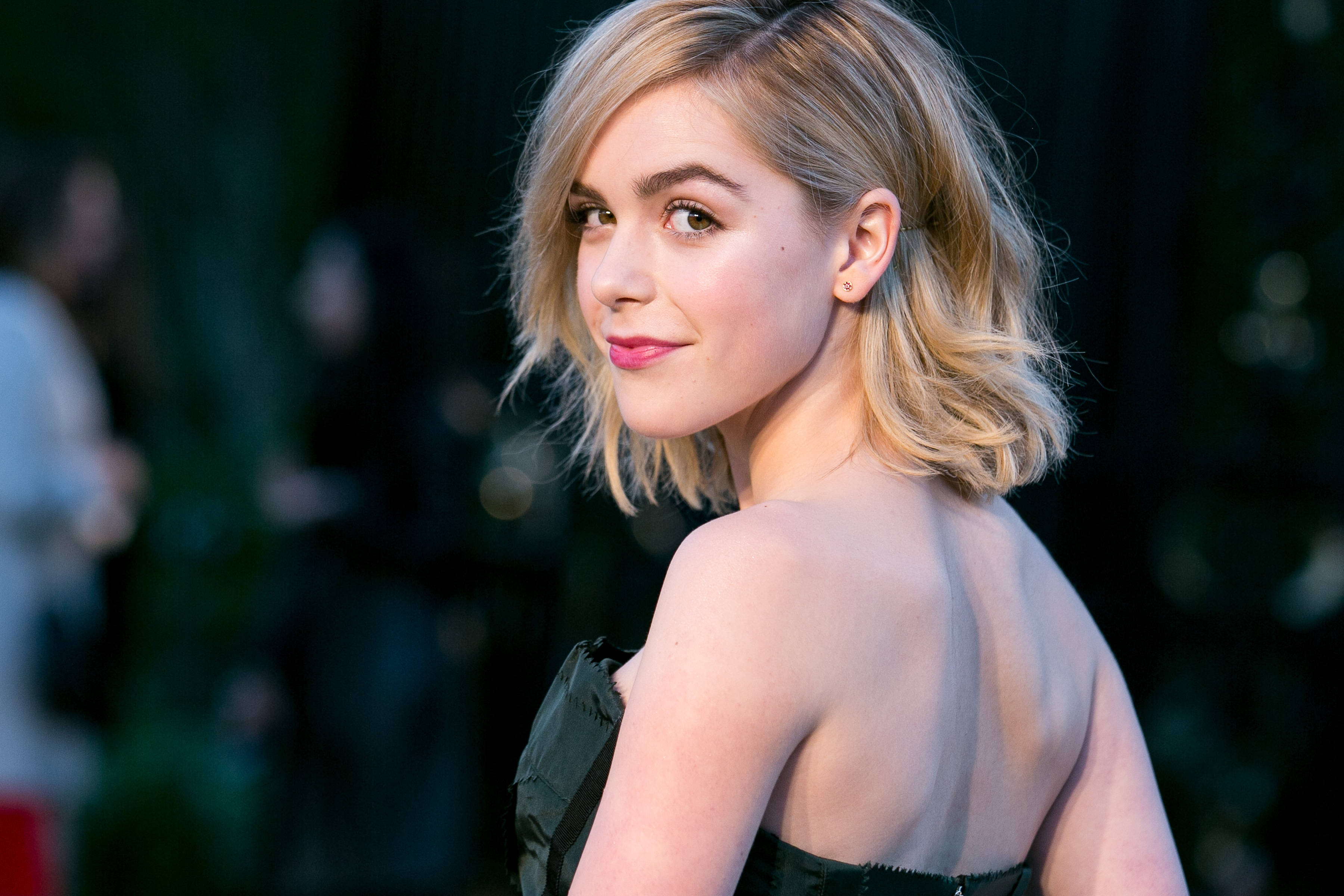 Kiernan Shipka 桌布 壁紙 — 一位金色鮑伯頭、粉紅嘴唇的女性，穿著深綠色無肩帶連衣裙，回頭微笑。