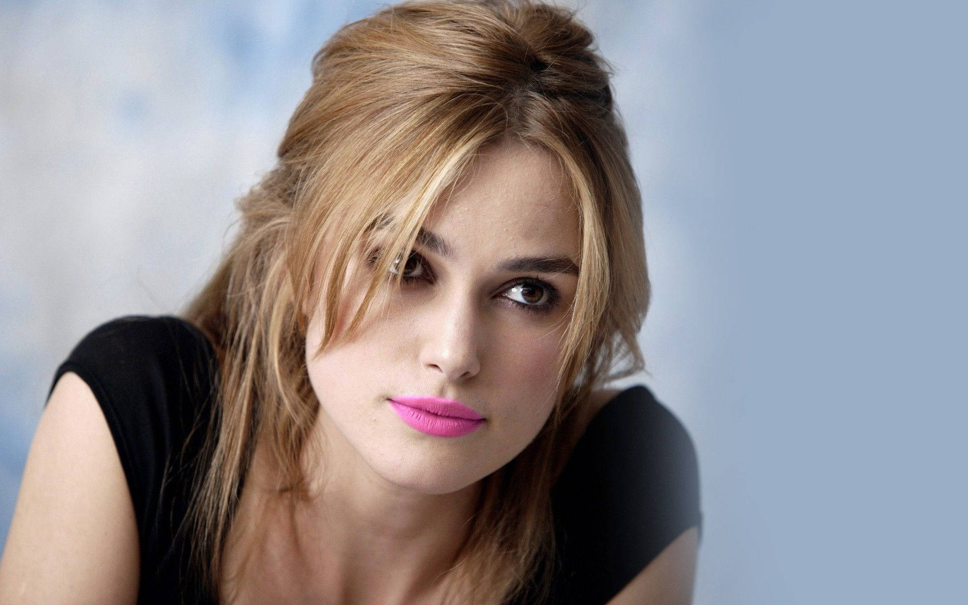Keira Knightley 桌布 壁紙 — 一位金髮、塗著大膽粉紅色唇膏的女性，在柔和的藍色背景下凝視著。