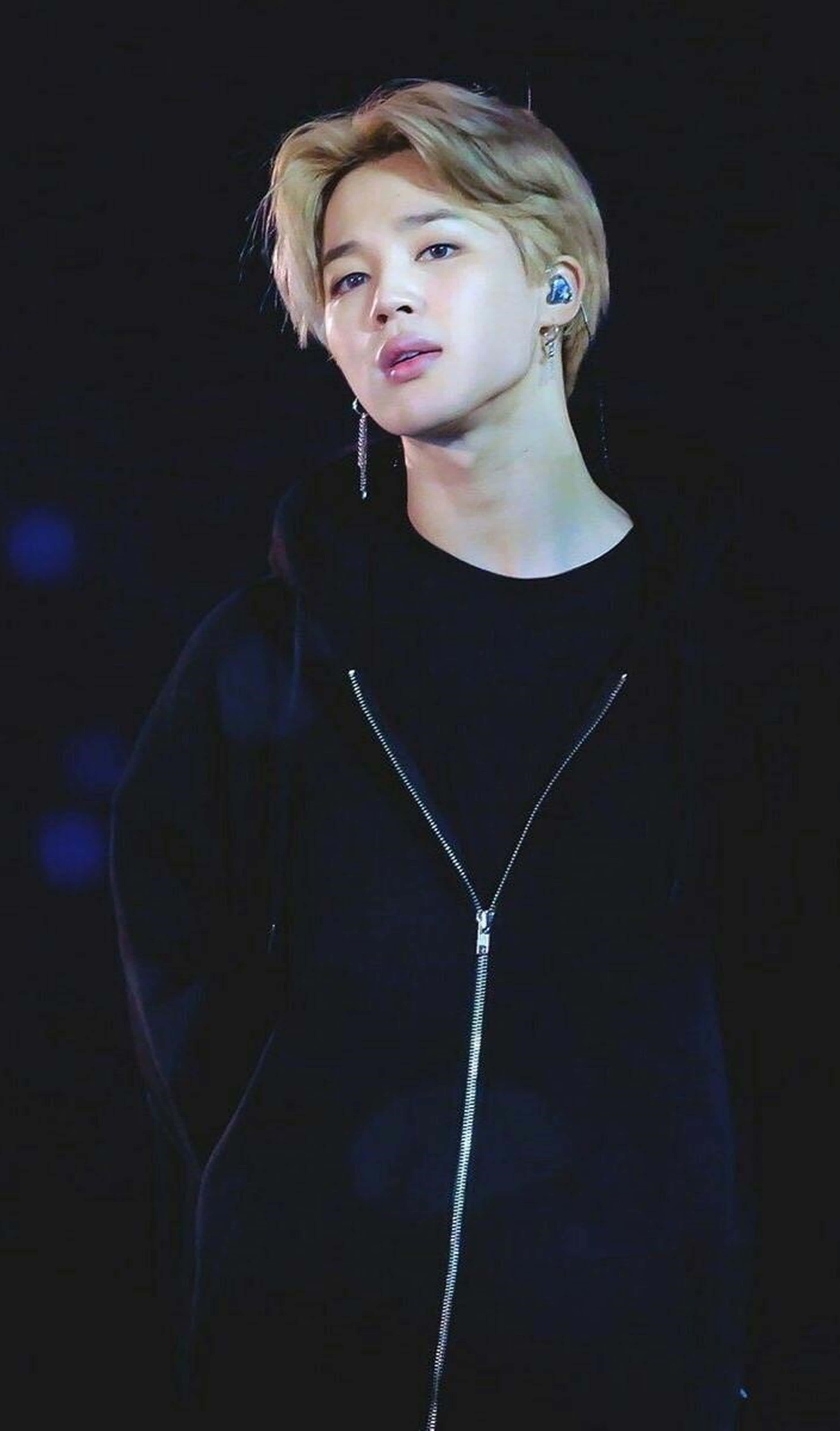 Bts Jimin 桌布 壁紙 — 一位金髮、戴銀耳環的年輕男子，在舞台燈光下穿著黑色拉鍊連帽衫，溫柔地凝視。