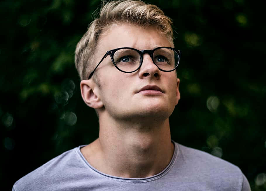 Boy 桌布 壁紙 — 一位金髮、戴黑框眼鏡的年輕男子，在模糊的綠色樹葉背景下向上凝視。