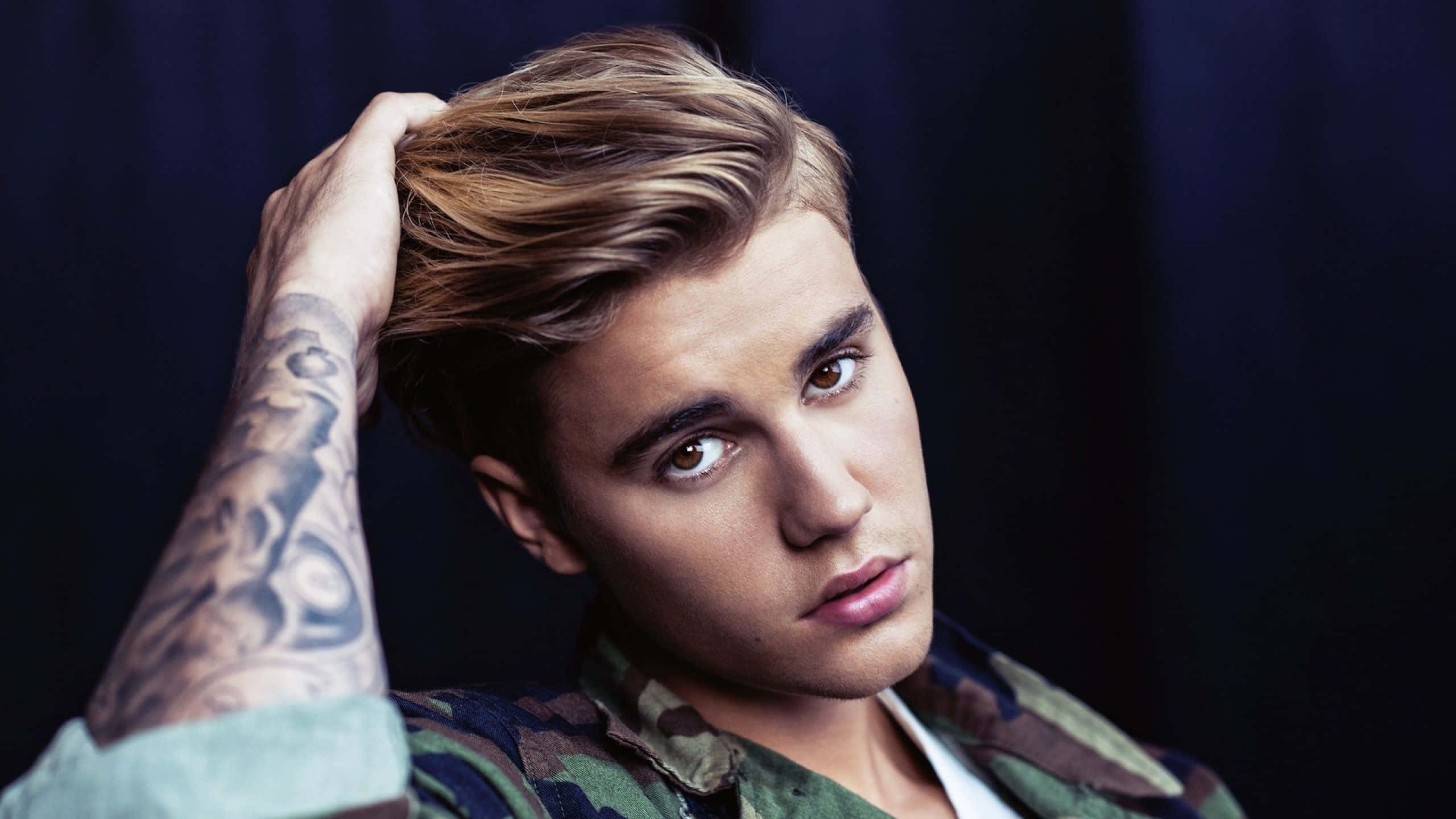 Justin Bieber 2021 桌布 壁紙 — 一位金髮、手臂有刺青的年輕男子，在深藍色背景下用手撥弄頭髮。
