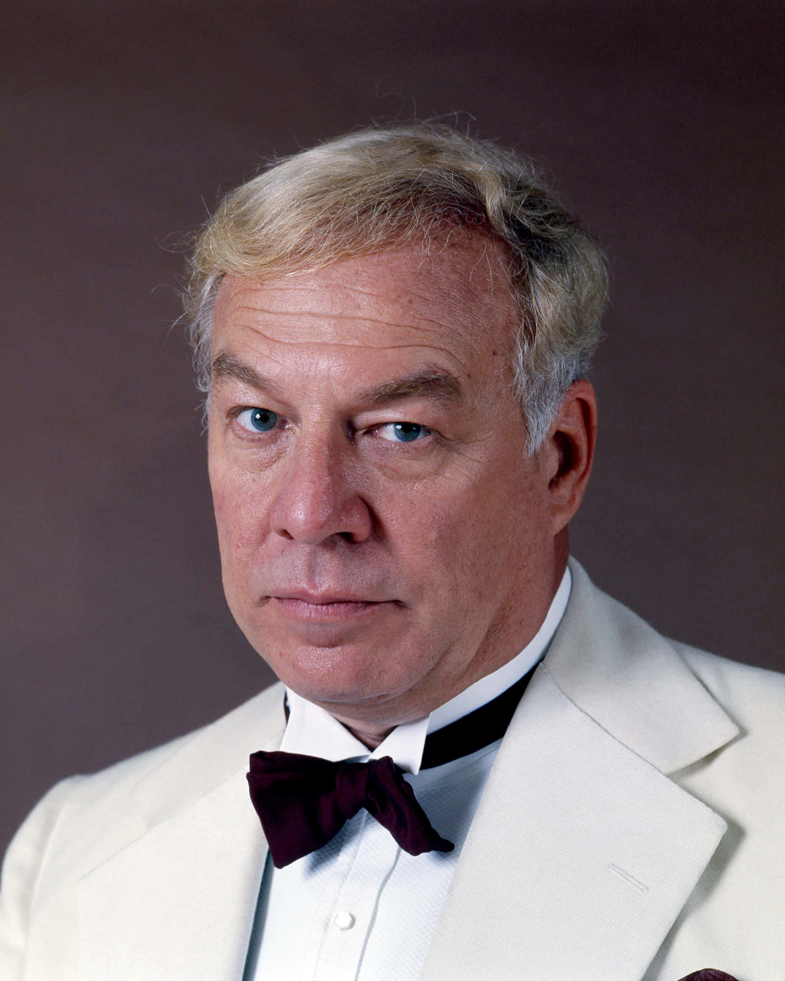 George Kennedy 桌布 壁紙 — 一位金髮、穿著白色燕尾服和酒紅色領結的尊貴長者，在柔和棕色背景下的特寫肖像。