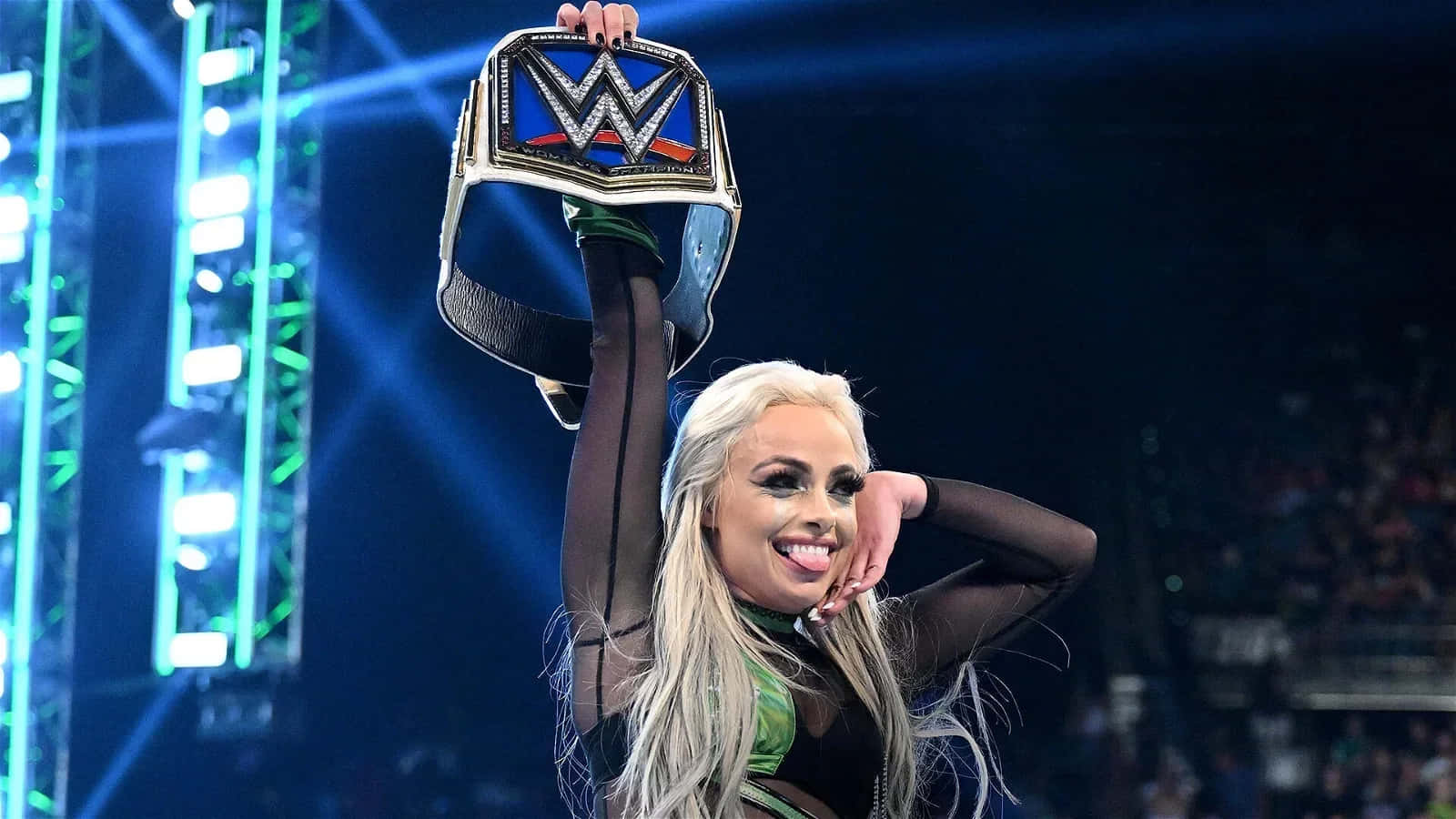 WWE 桌布 壁紙 — 一位金髮WWE摔角手在擠滿觀眾的場館中，於明亮的舞台燈光下勝利地高舉冠軍腰帶。