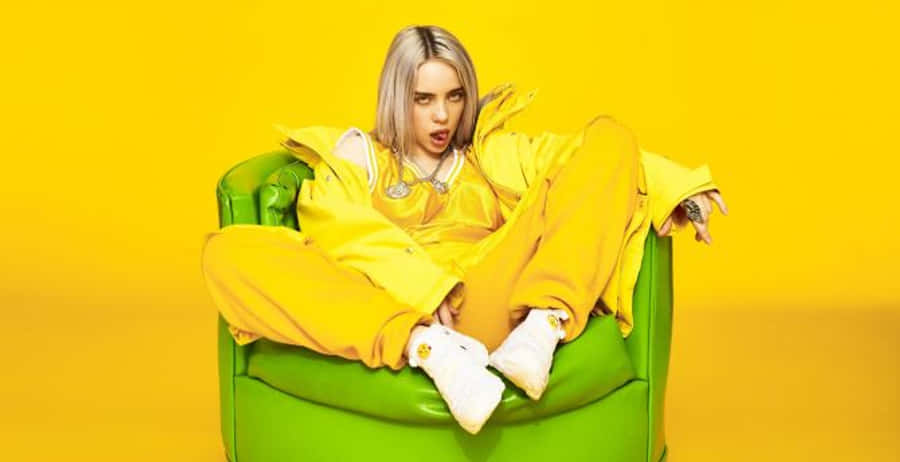 Billie Eilish Laptop 桌布 壁紙 — 一位金髮人士休閒地坐在綠色扶手椅上，身穿亮黃色服裝，背景為純黃色。