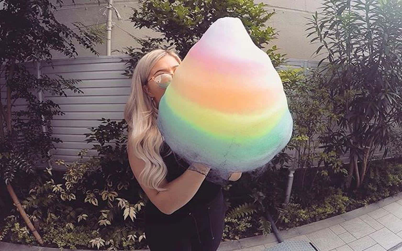 Cotton Candy 桌布 壁紙 — 一位金髮人士在戶外靠近植物和圍欄處手持巨型彩虹棉花糖。
