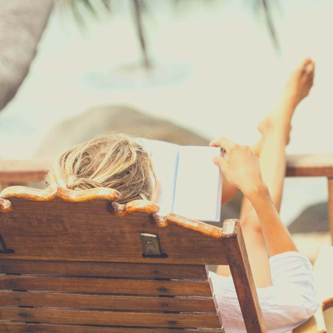 Contentment 桌布 壁紙 — 一位金髮人士在木製躺椅上放鬆，在海邊棕櫚樹下閱讀書籍。
