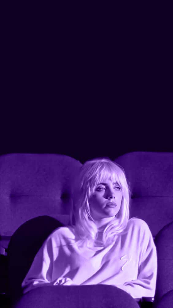 Billie Eilish Purple 桌布 壁紙 — 一位金髮人士坐在昏暗的劇院座位上，沐浴在紫色燈光中，若有所思地向前凝視。