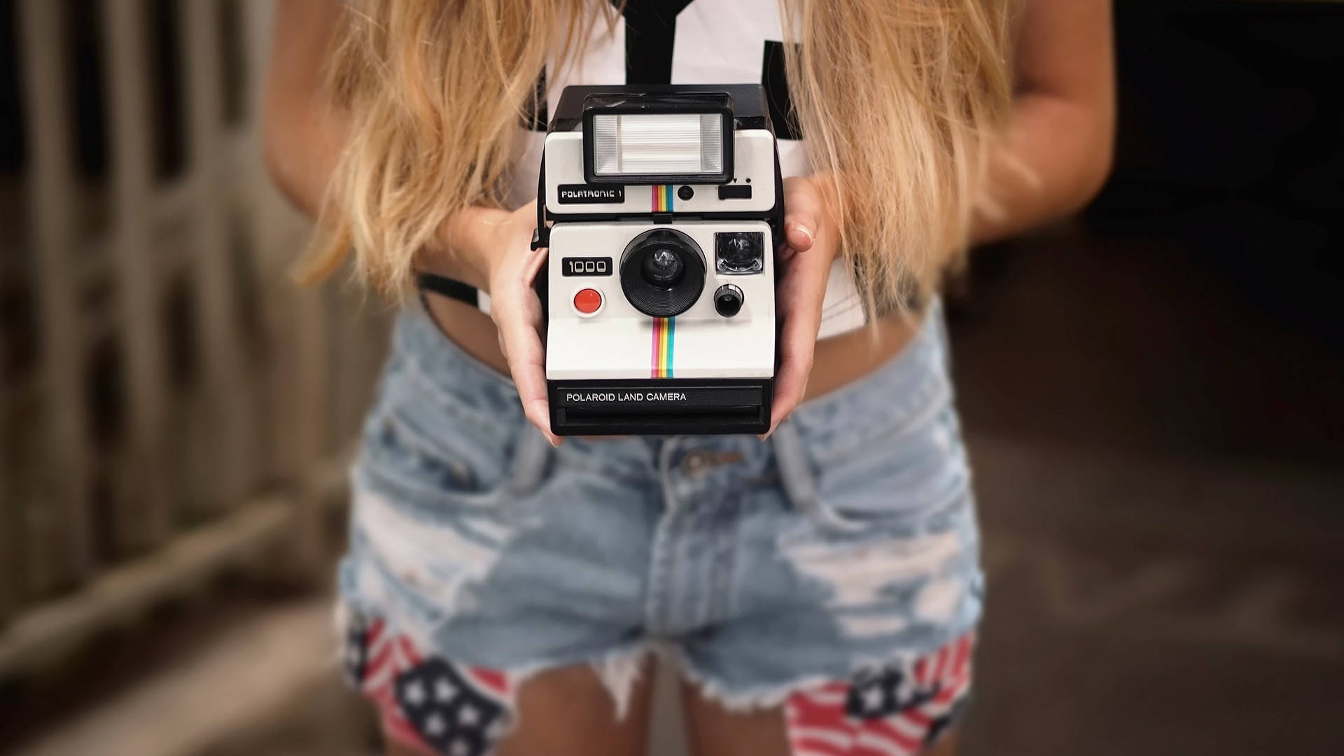 拍立得 桌布 壁紙 — 一位金髮人士手持復古Polaroid Land Camera，身穿帶有美國國旗補丁的破損牛仔短褲。