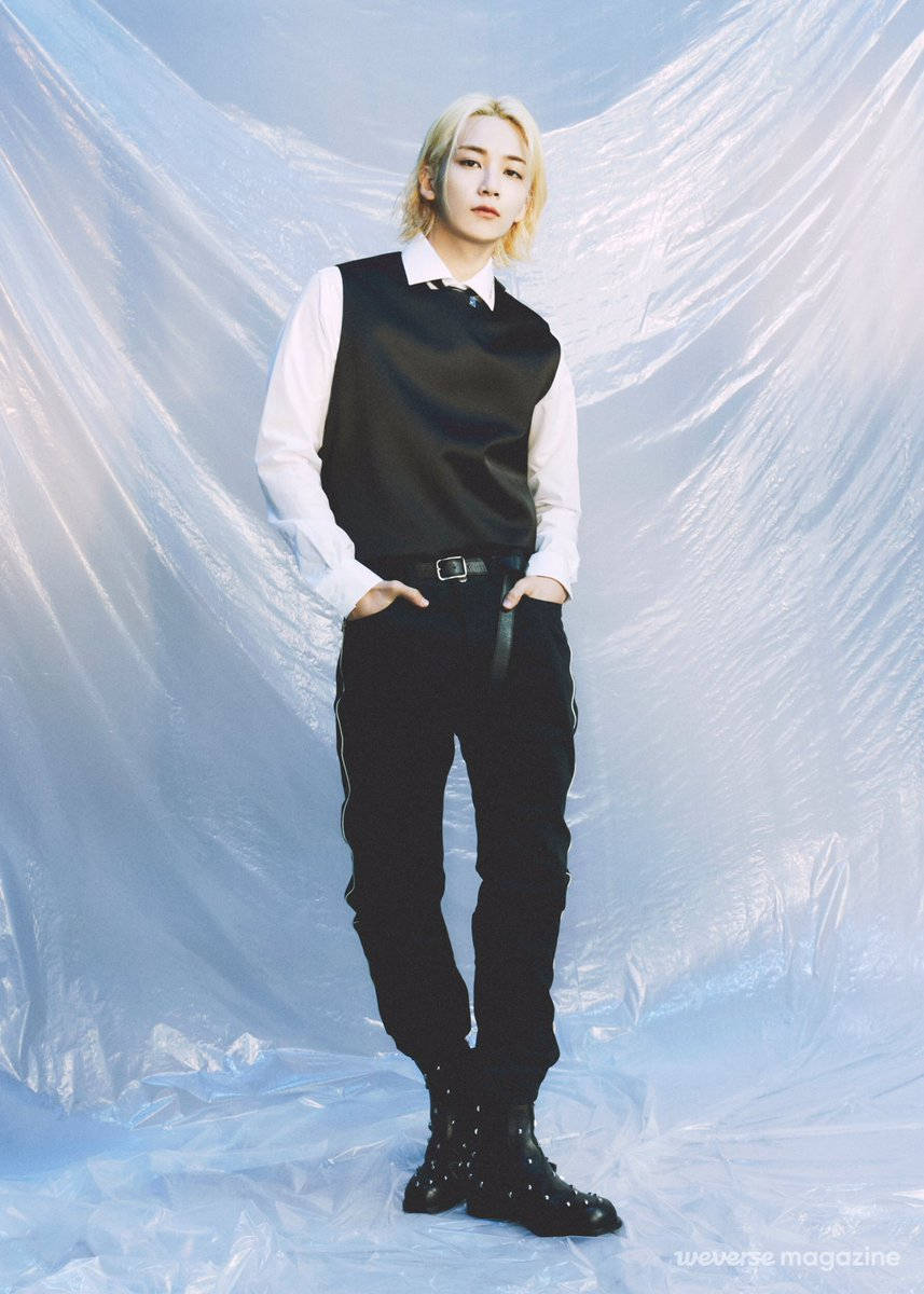 yoon jeonghan 桌布 壁紙 — 一位金髮人士自信地穿著黑色背心和褲子，站在皺褶的半透明藍色背景前。