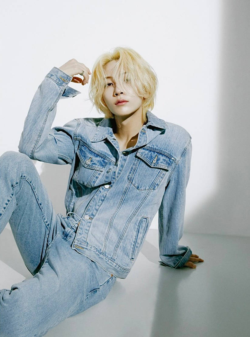 yoon jeonghan 桌布 壁紙 — 一位金髮人物穿著牛仔服裝，在純白背景和戲劇性燈光下隨意坐著。