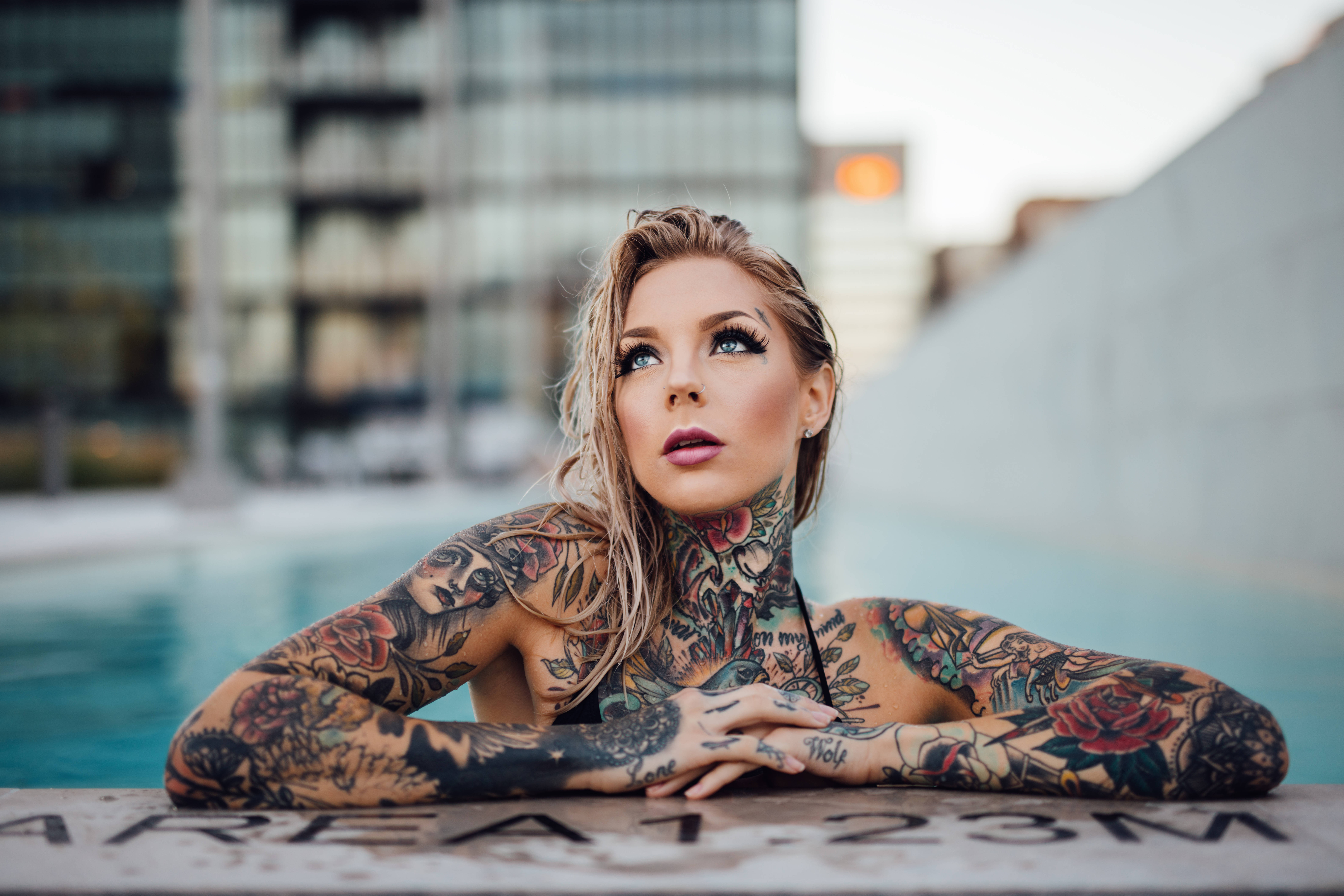 Tattoo 桌布 壁紙 — 一位金髮刺青女子在黃昏時分，手臂擱在泳池邊，仰望模糊的城市背景。