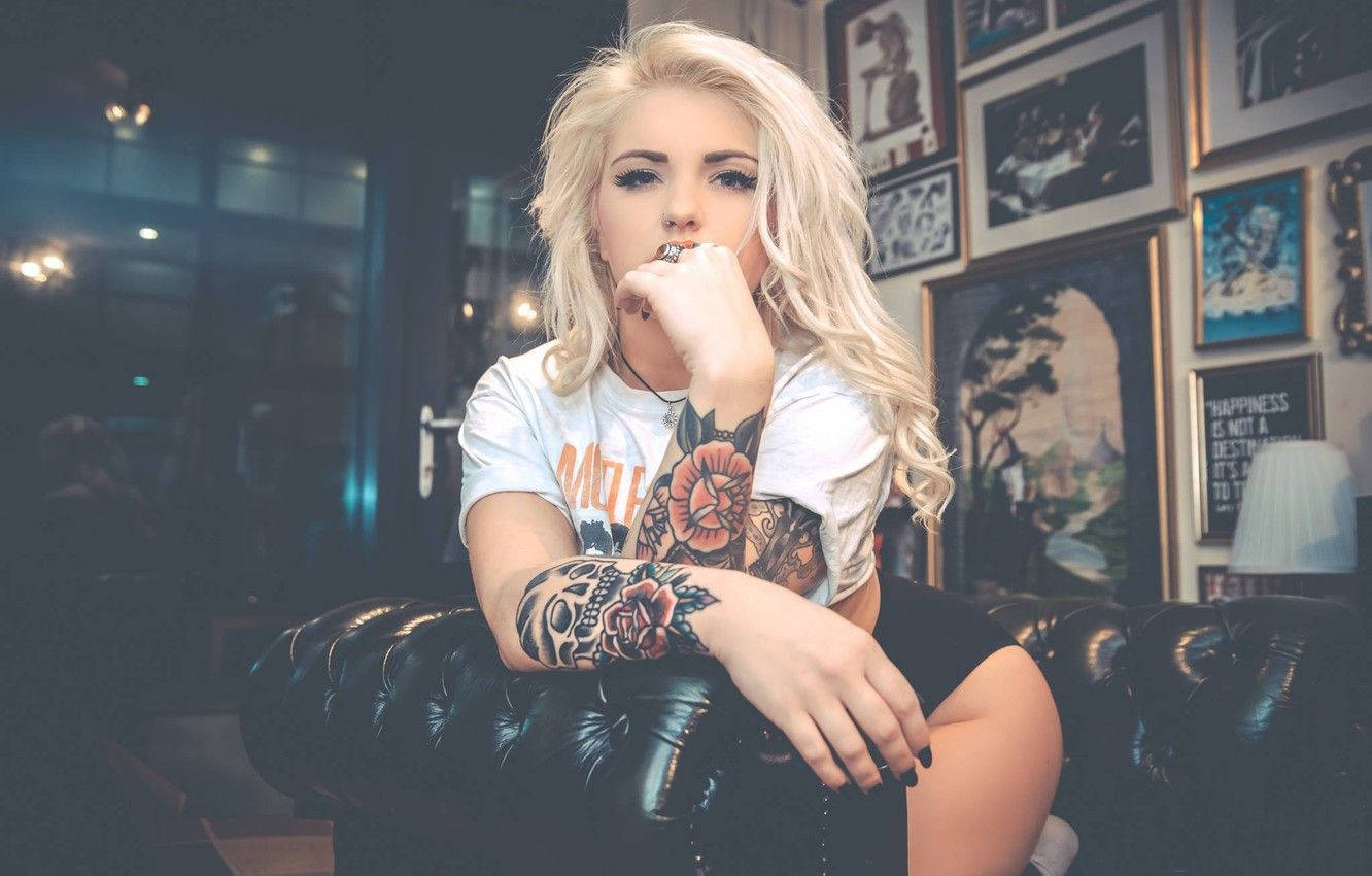 tattoos 桌布 壁紙 — 一位金髮刺青女子自信地坐在黑色皮沙發上，房間燈光昏暗，裝飾著裱框藝術品。