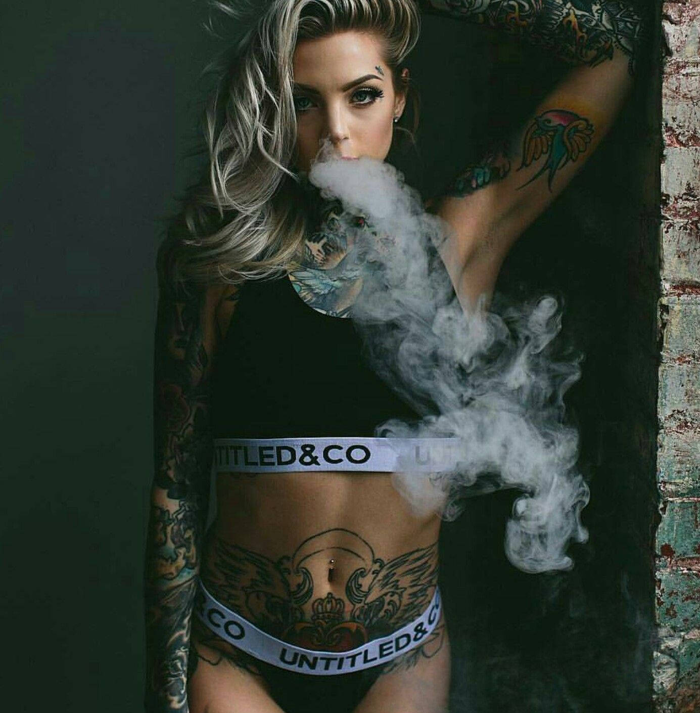 hot girl 桌布 壁紙 — 一位金髮刺青女性在深色紋理牆壁前穿著品牌內衣呼出煙霧。