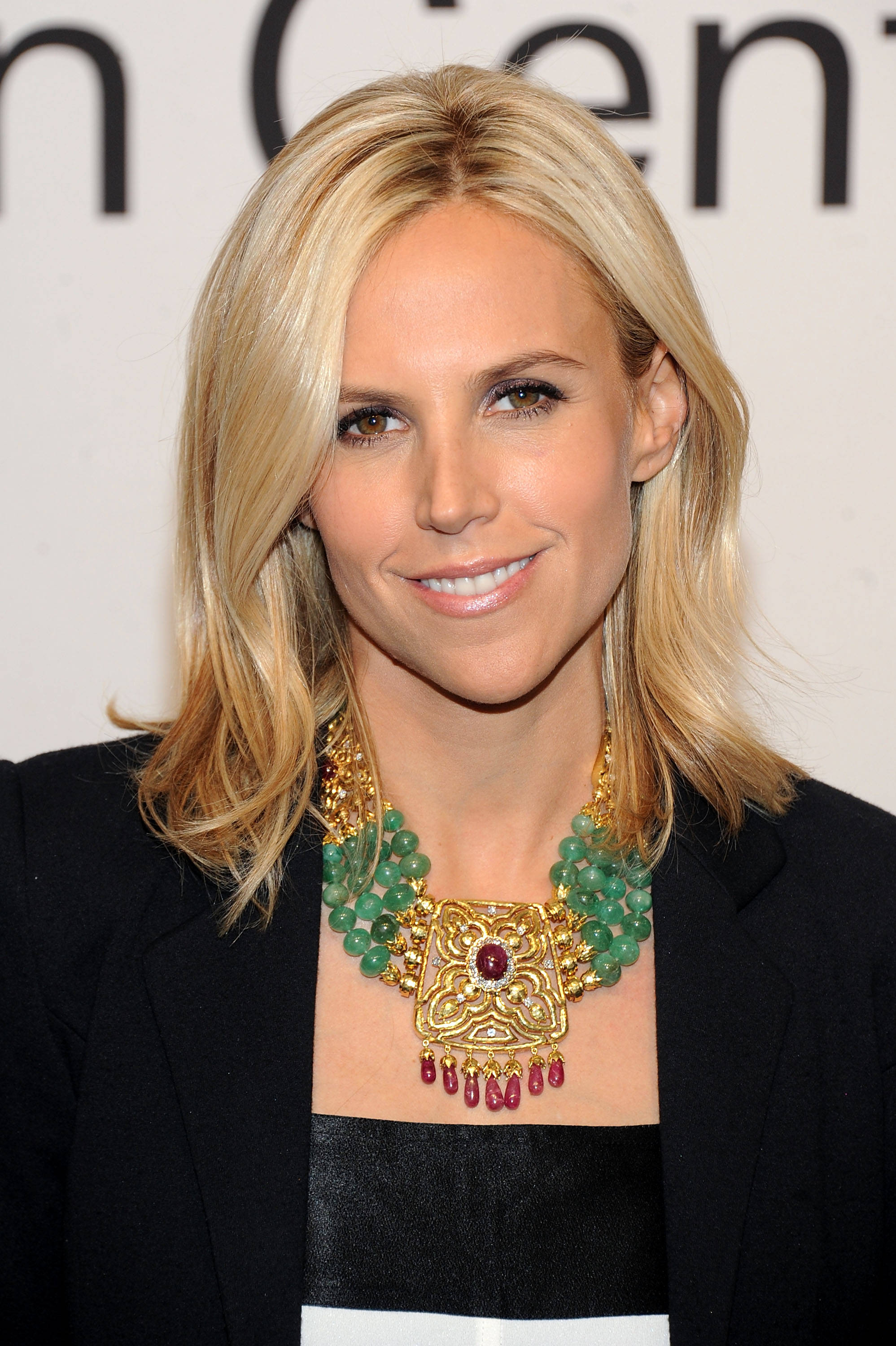 Tory Burch 桌布 壁紙 — 一位金髮女士在正式活動中微笑，佩戴著大膽的金色和綠色寶石項鍊。