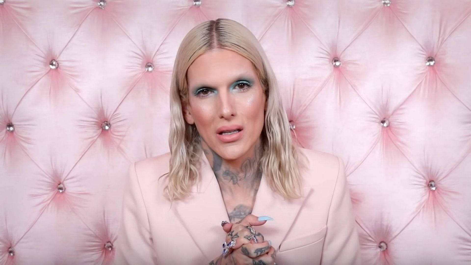 Jeffree Star 桌布 壁紙 — 一位金髮女子，帶有刺青和誇張妝容，坐在飾有水晶鈕扣的粉紅色簇絨牆前。