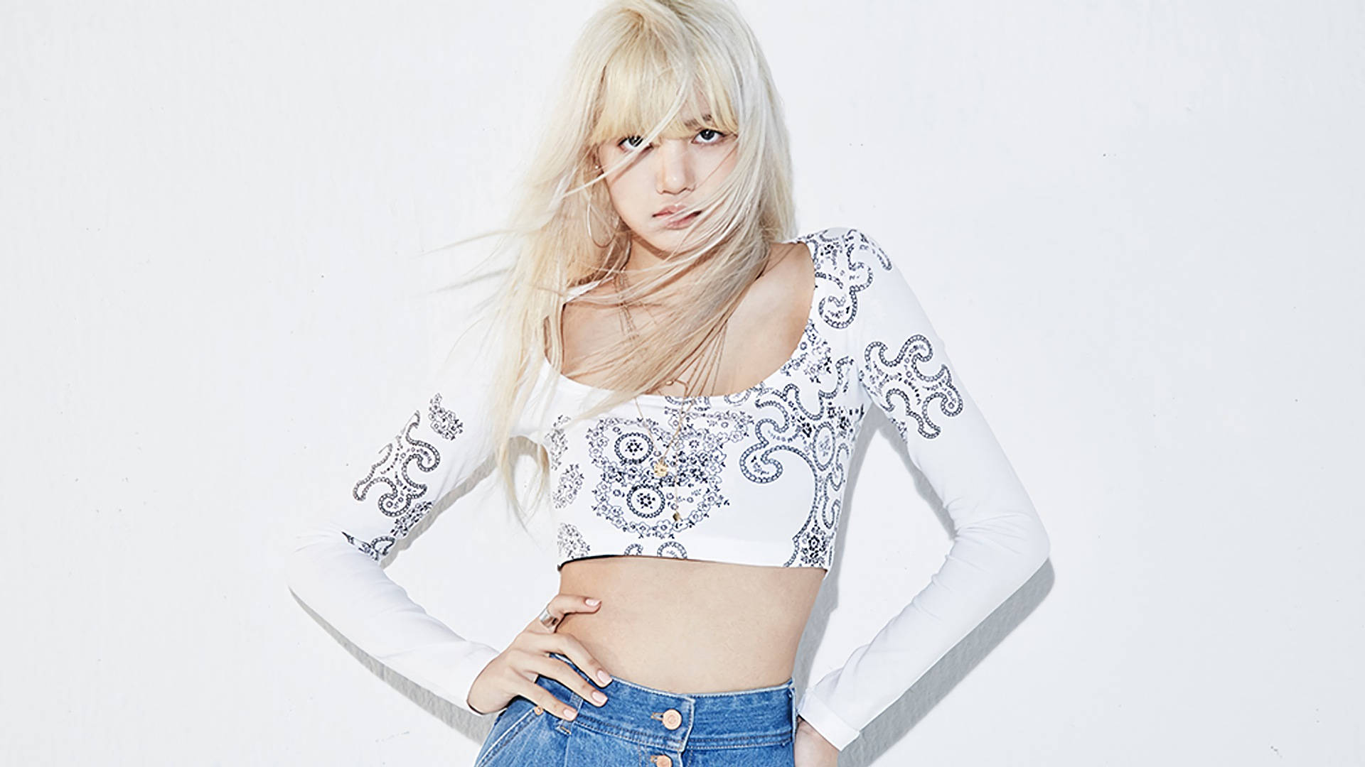 Lalisa 桌布 壁紙 — 一位金髮女子，頭髮隨風飄揚，穿著白色圖案露臍上衣和牛仔褲，在純白背景前自信地擺姿勢。