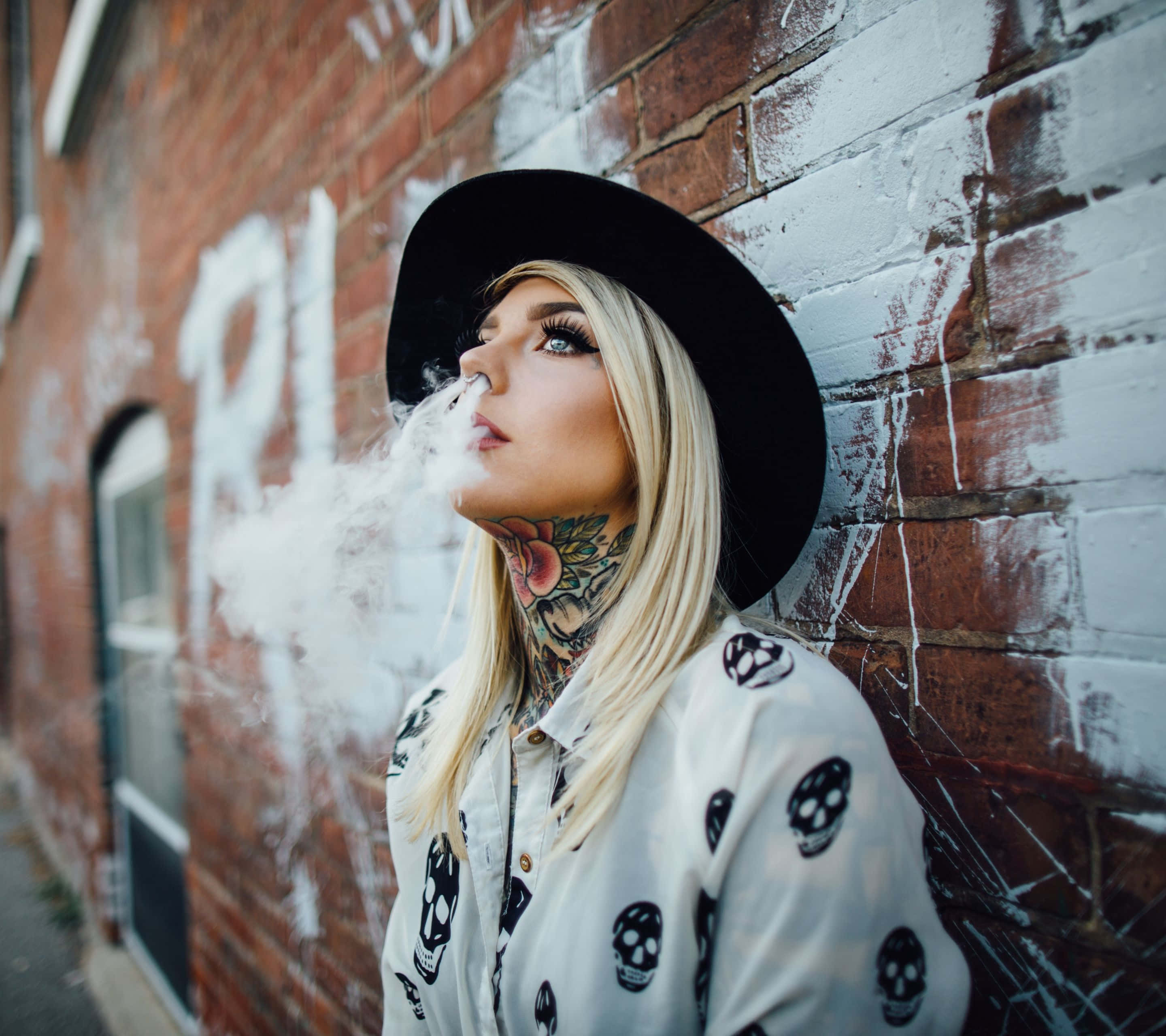 Tattoo 桌布 壁紙 — 一位金髮女子，頸部有刺青，戴著黑色帽子，靠著塗鴉磚牆呼出煙霧。