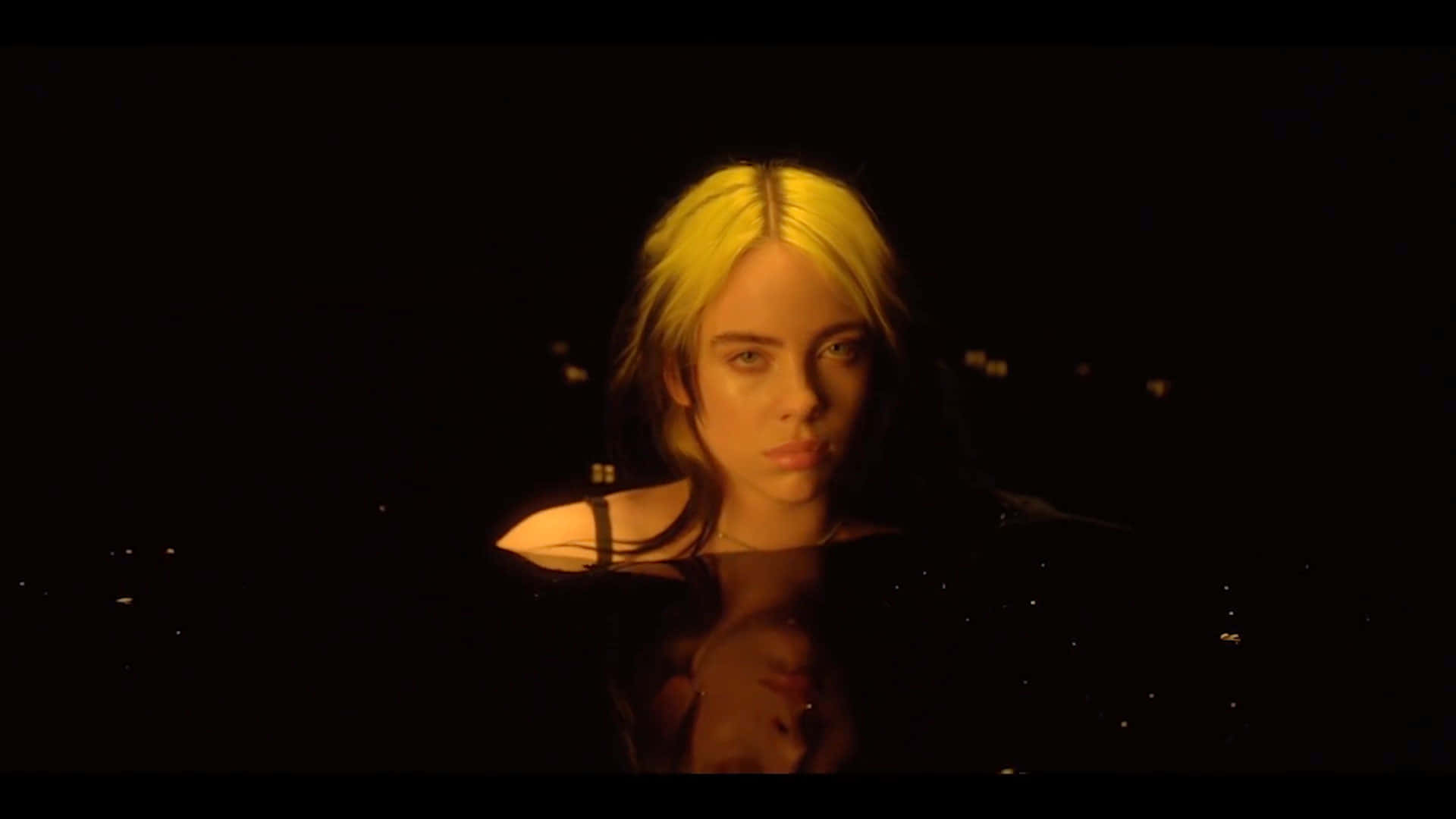 Billie Eilish Laptop 桌布 壁紙 — 一位金髮女子在溫暖的金色燈光下，從黑暗的水面專注地凝視。