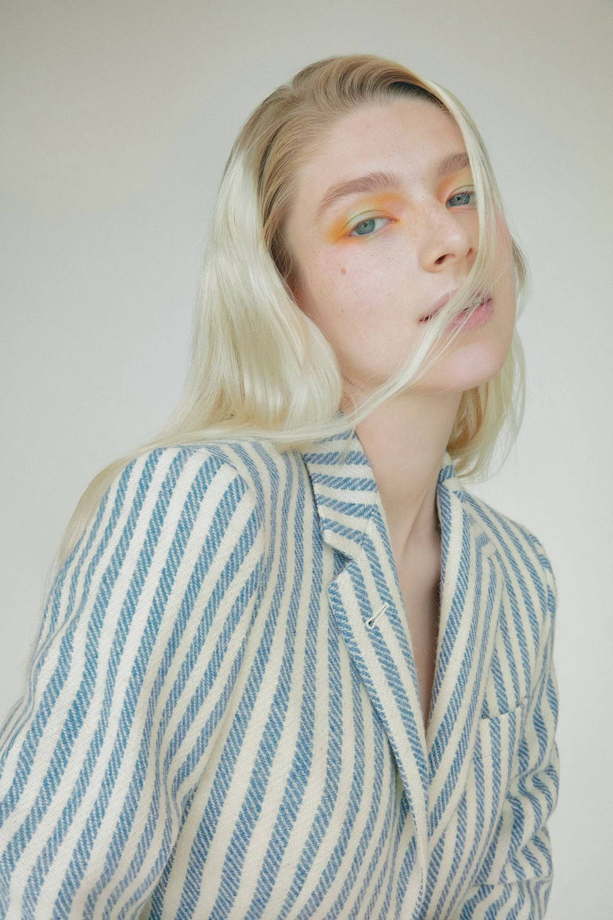 Hunter Schafer 桌布 壁紙 — 一位金髮女子塗上搶眼的橙色眼影，一縷髮絲飄過臉龐，在純色背景前溫柔凝視。