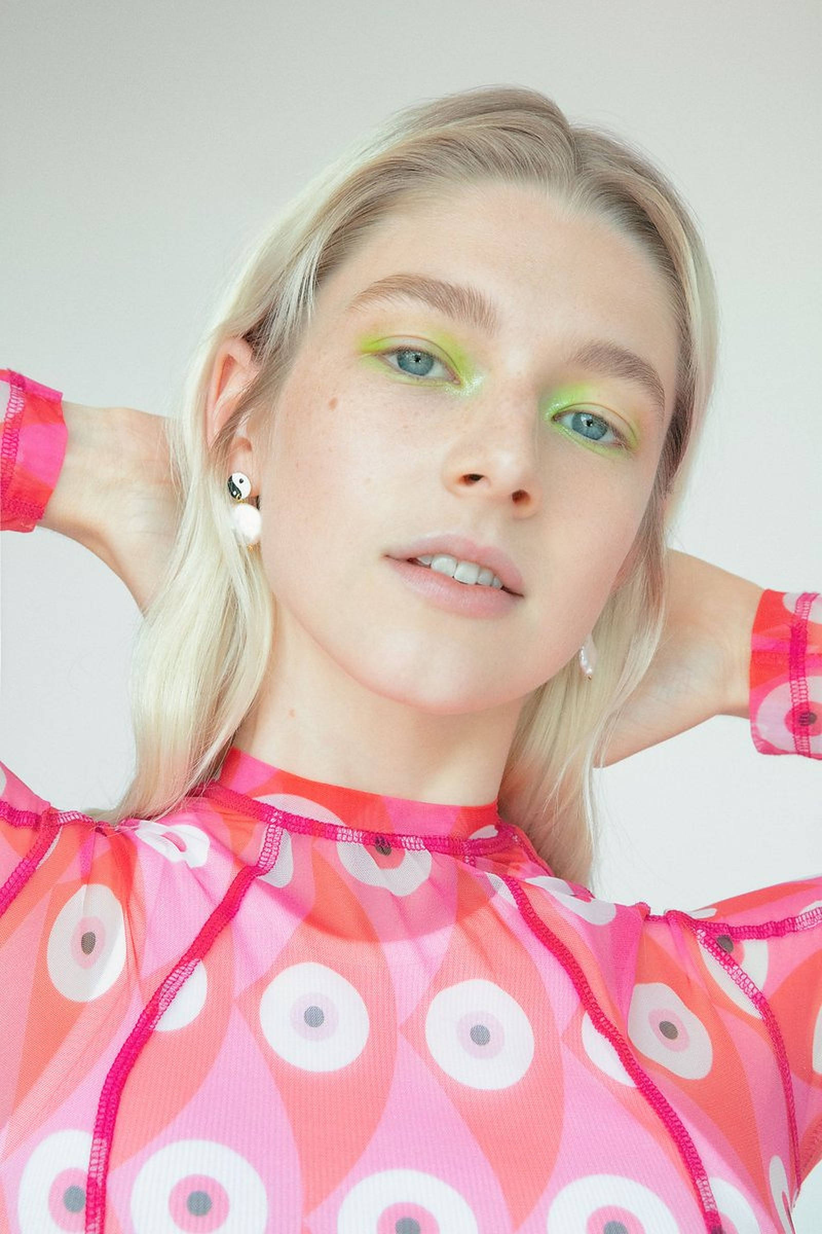 Hunter Schafer 桌布 壁紙 — 一位金髮女子塗上螢光綠色眼影，身穿帶有白色圓形圖案的透視粉紅色上衣，背景為純色。