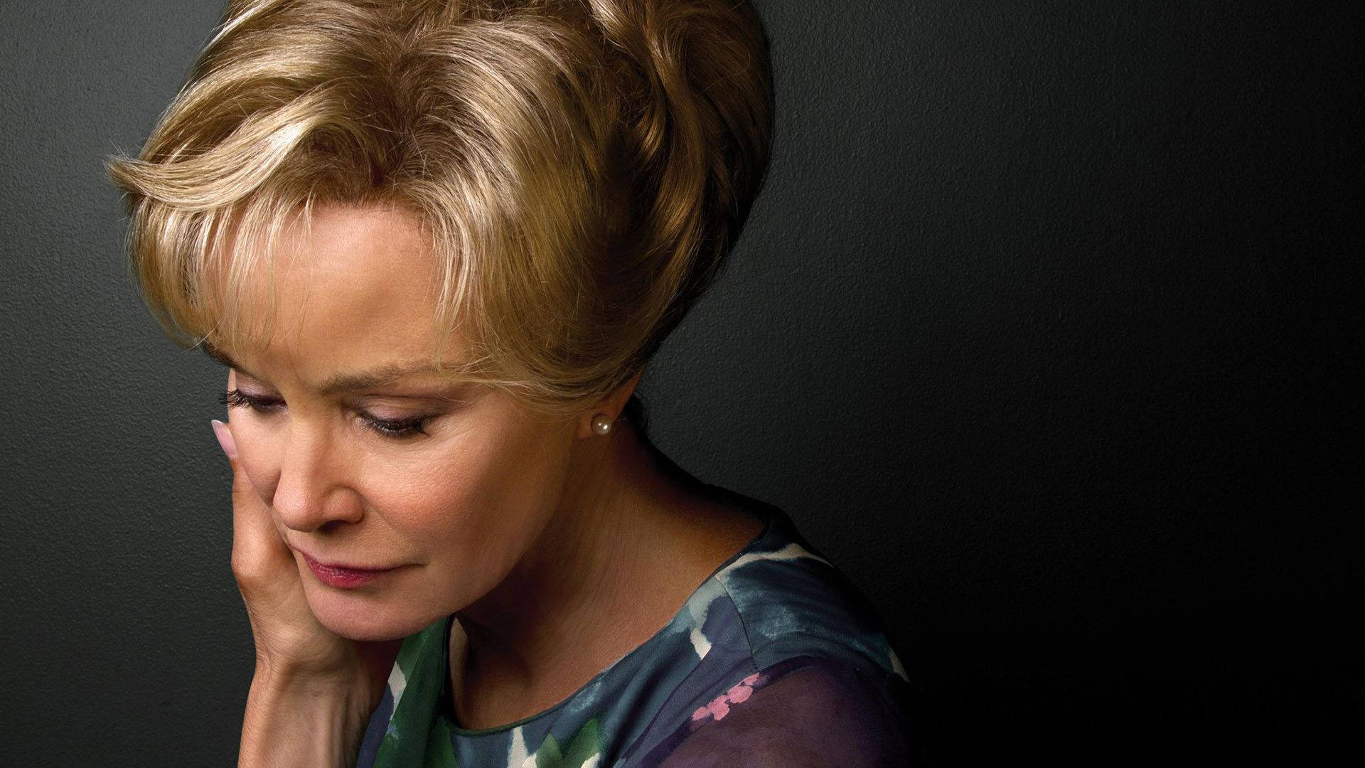 Jessica Lange 桌布 壁紙 — 一位金髮女子將頭靠在手上，倚著深色紋理牆壁，雙眼低垂，靜靜沉思。