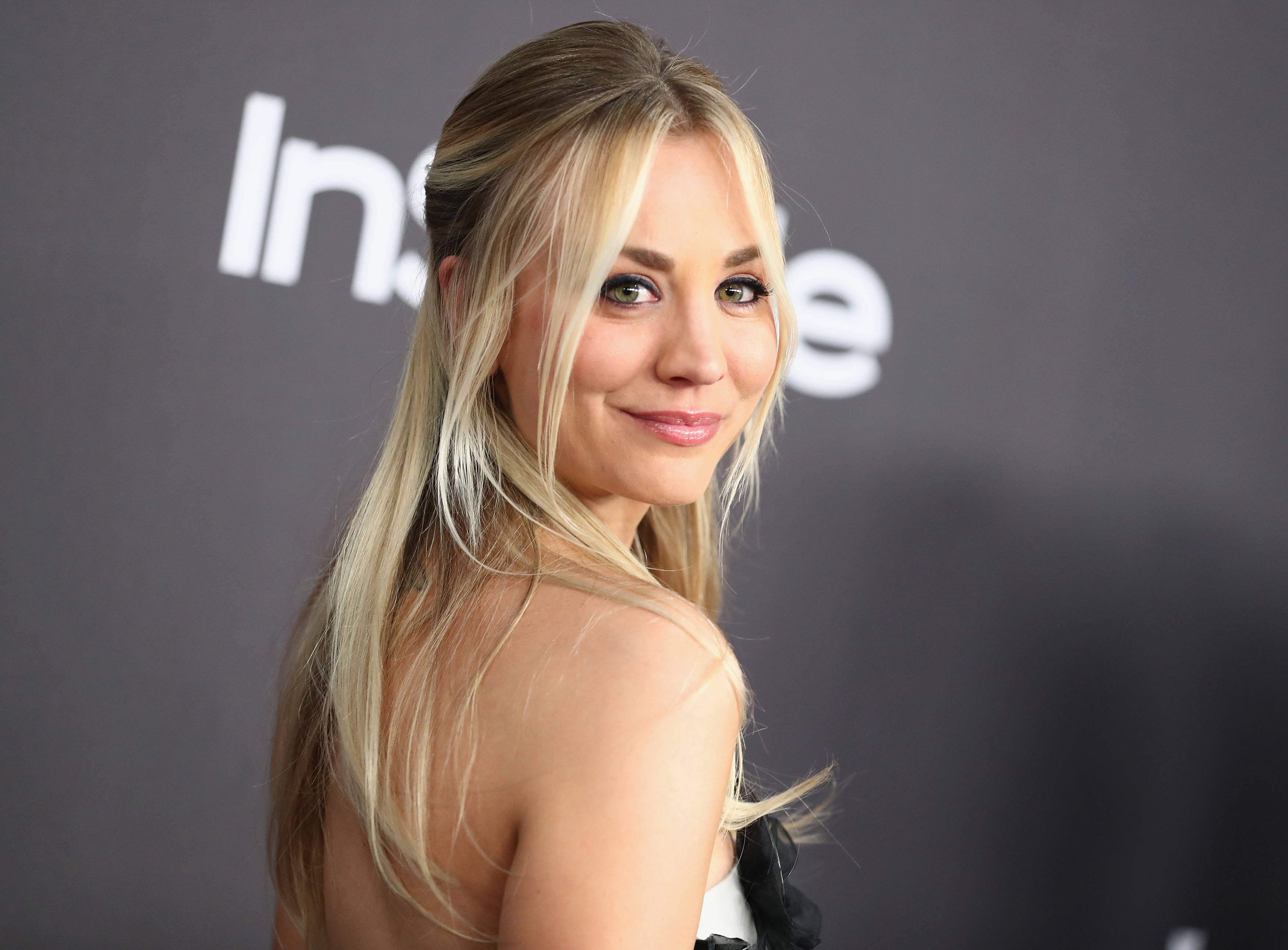 Kaley Cuoco 桌布 壁紙 — 一位金髮女子帶著溫柔的微笑，在深灰色背景上白色文字的襯托下，轉頭越過肩膀。