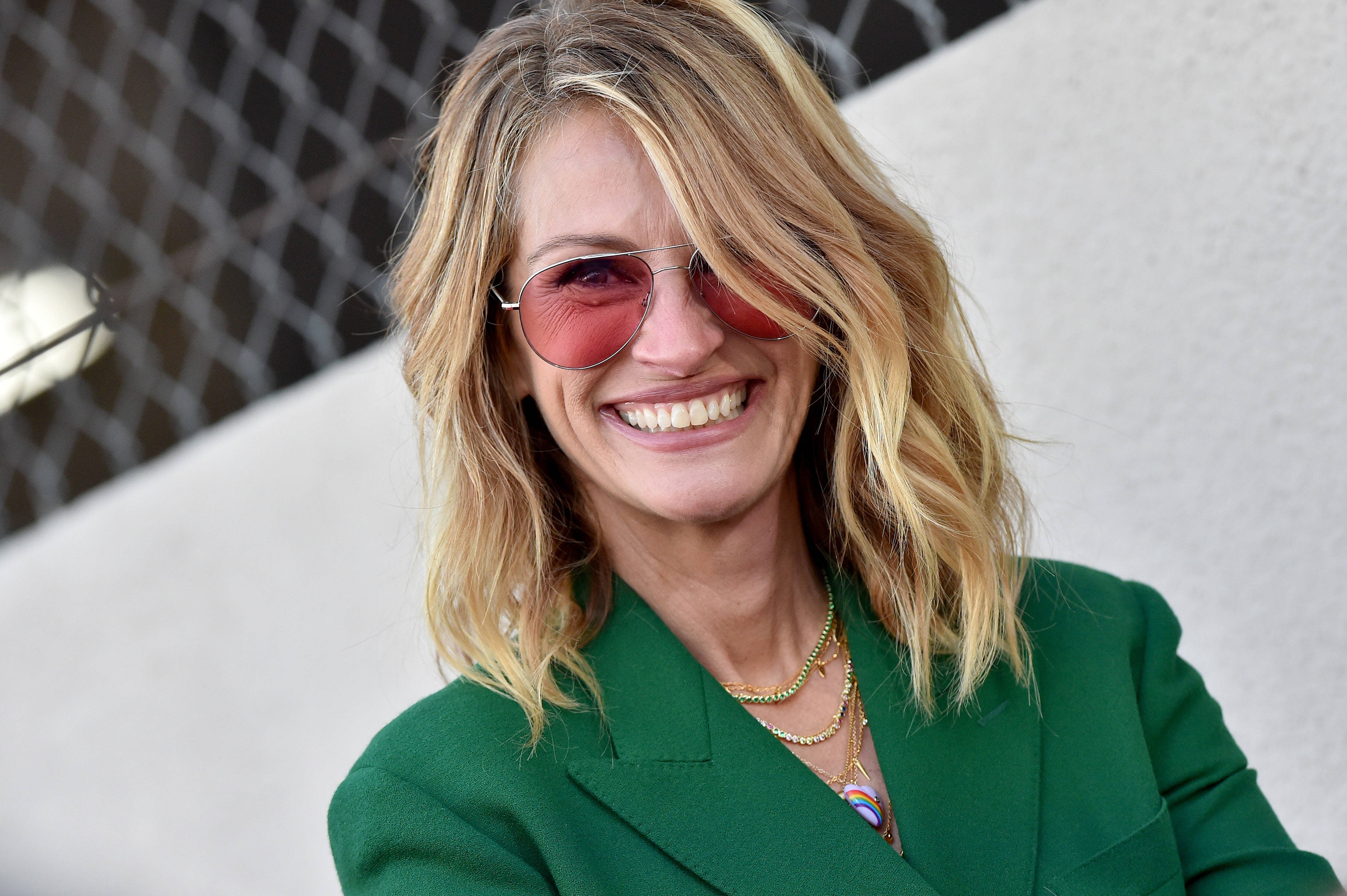 Julia Roberts 桌布 壁紙 — 一位金髮女子戴著紅色太陽眼鏡，身穿綠色西裝外套，在鐵絲網圍欄和白色牆壁前燦爛微笑。