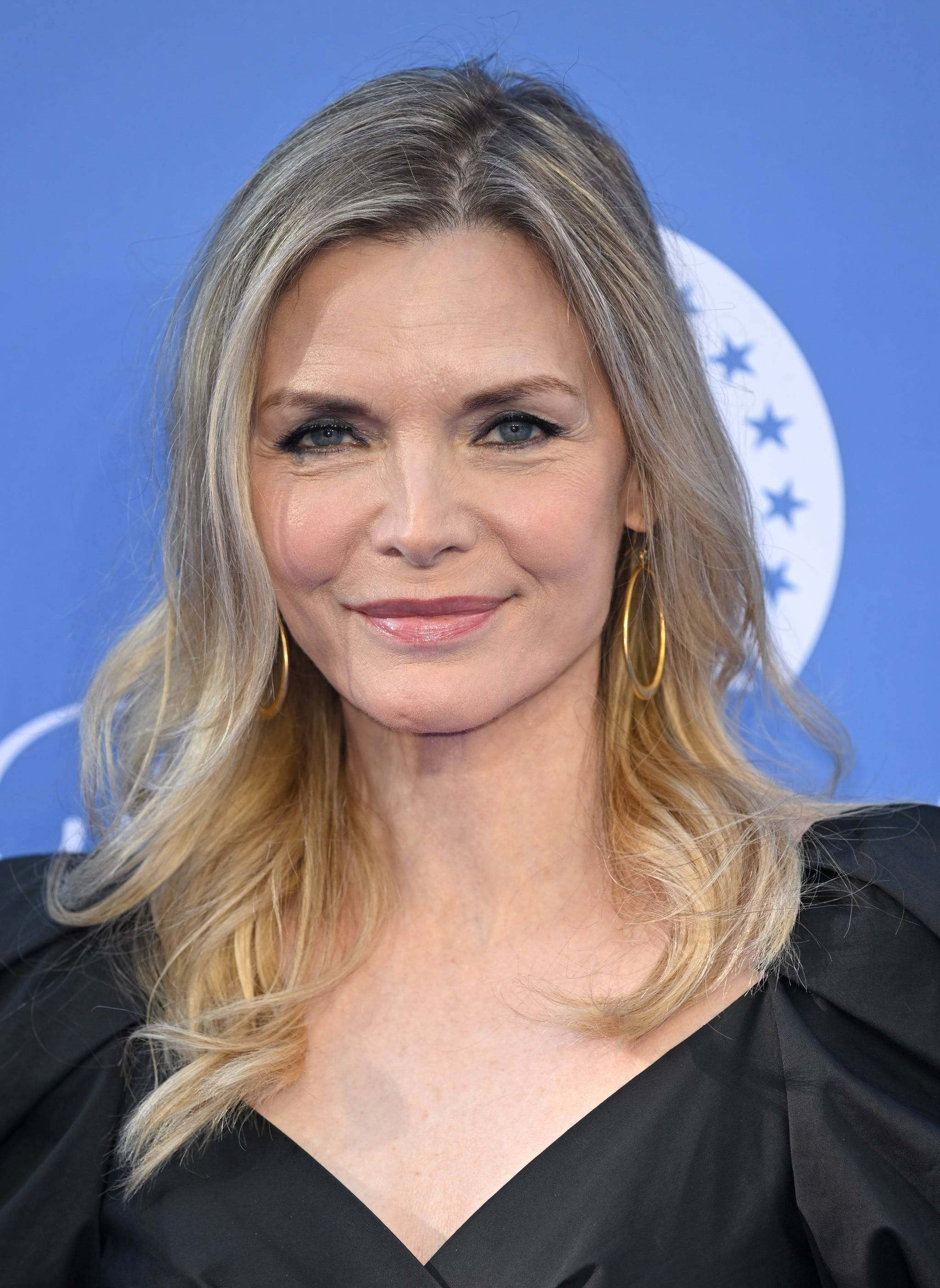 Celebrity 桌布 壁紙 — 一位金髮女子戴著金色圓環耳環，在帶有白色圓形標誌的藍色背景前微笑的特寫肖像。