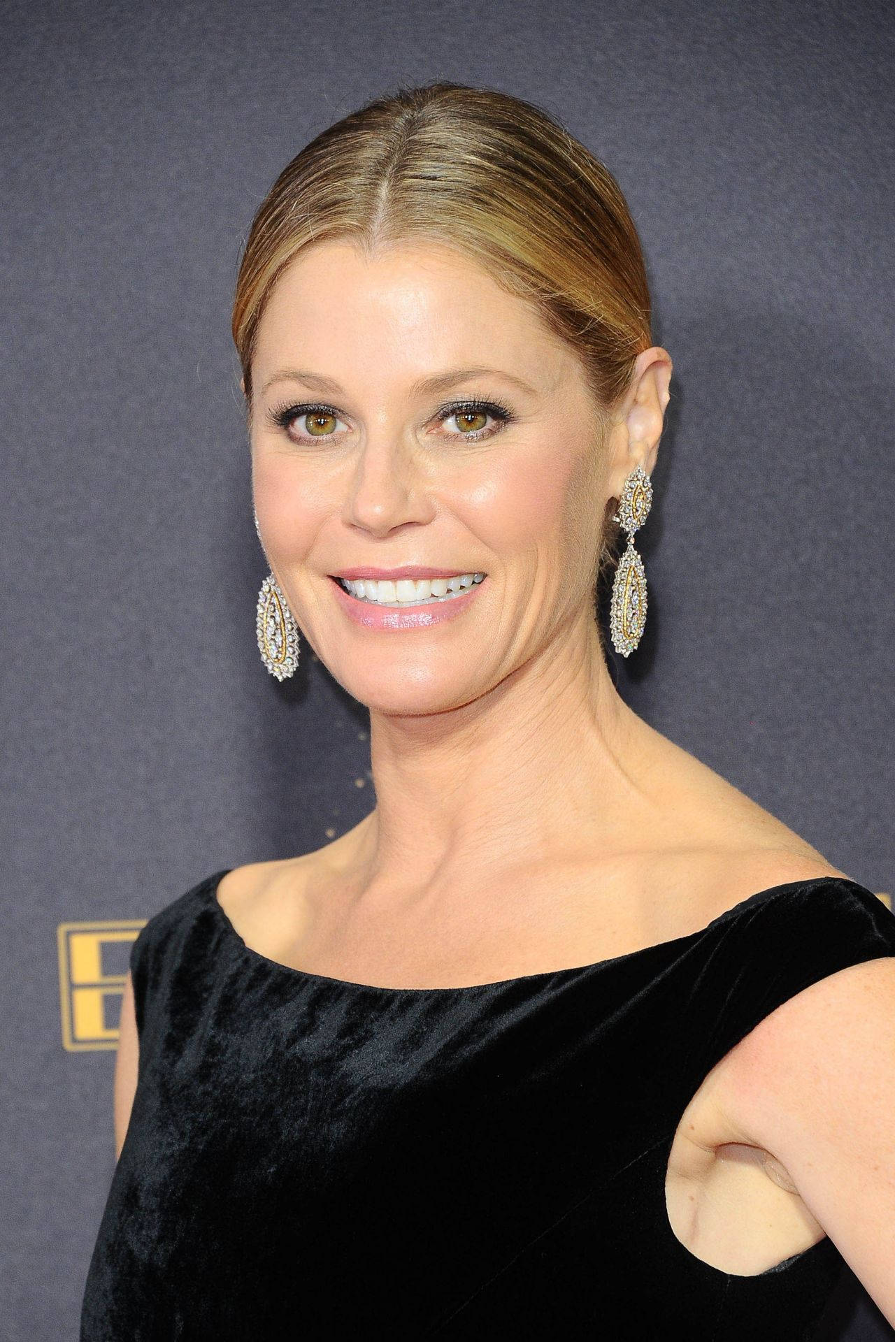 Julie Bowen 桌布 壁紙 — 一位金髮女子戴著閃亮耳環，身穿黑色天鵝絨連衣裙，在深色背景下溫暖微笑。