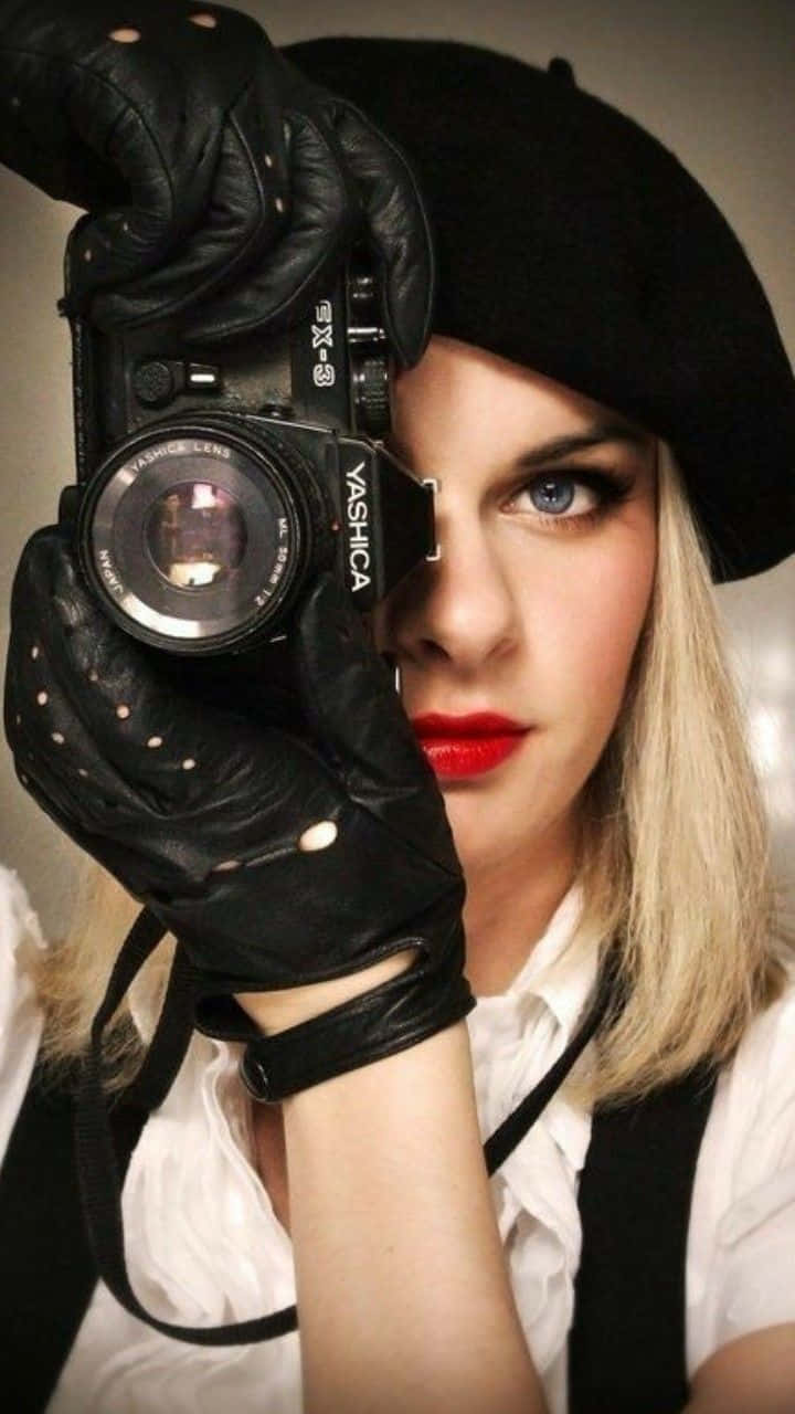 Holding Camera 桌布 壁紙 — 一位金髮女子戴著黑色貝雷帽和皮手套，將一部復古Yashica相機舉到眼前，擁有強烈的藍色眼睛和鮮豔的紅唇。