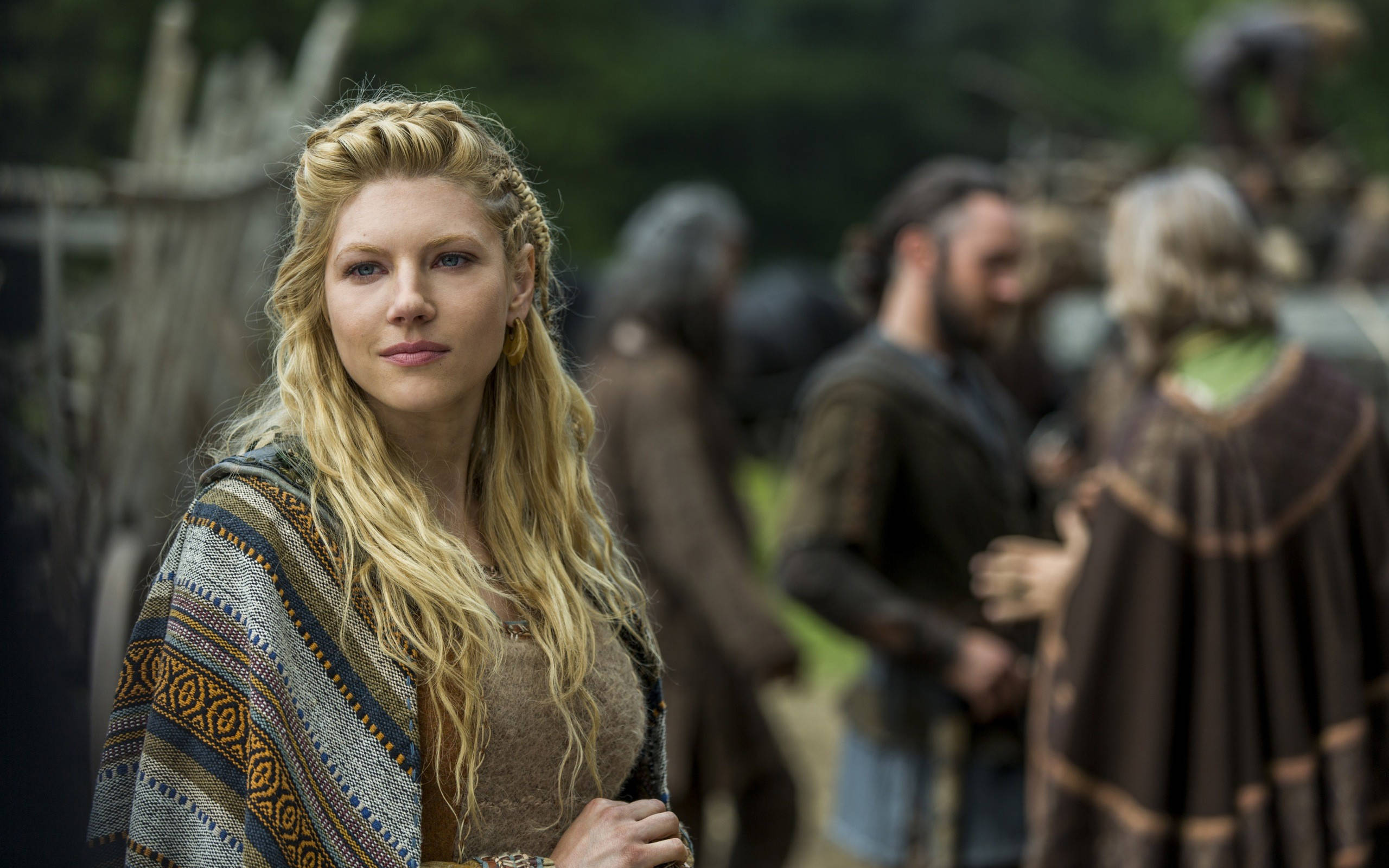 Vikings 桌布 壁紙 — 一位金髮女子披著圖案披肩，神情嚴肅地向前凝視，身後模糊的人影在森林環境中交錯。