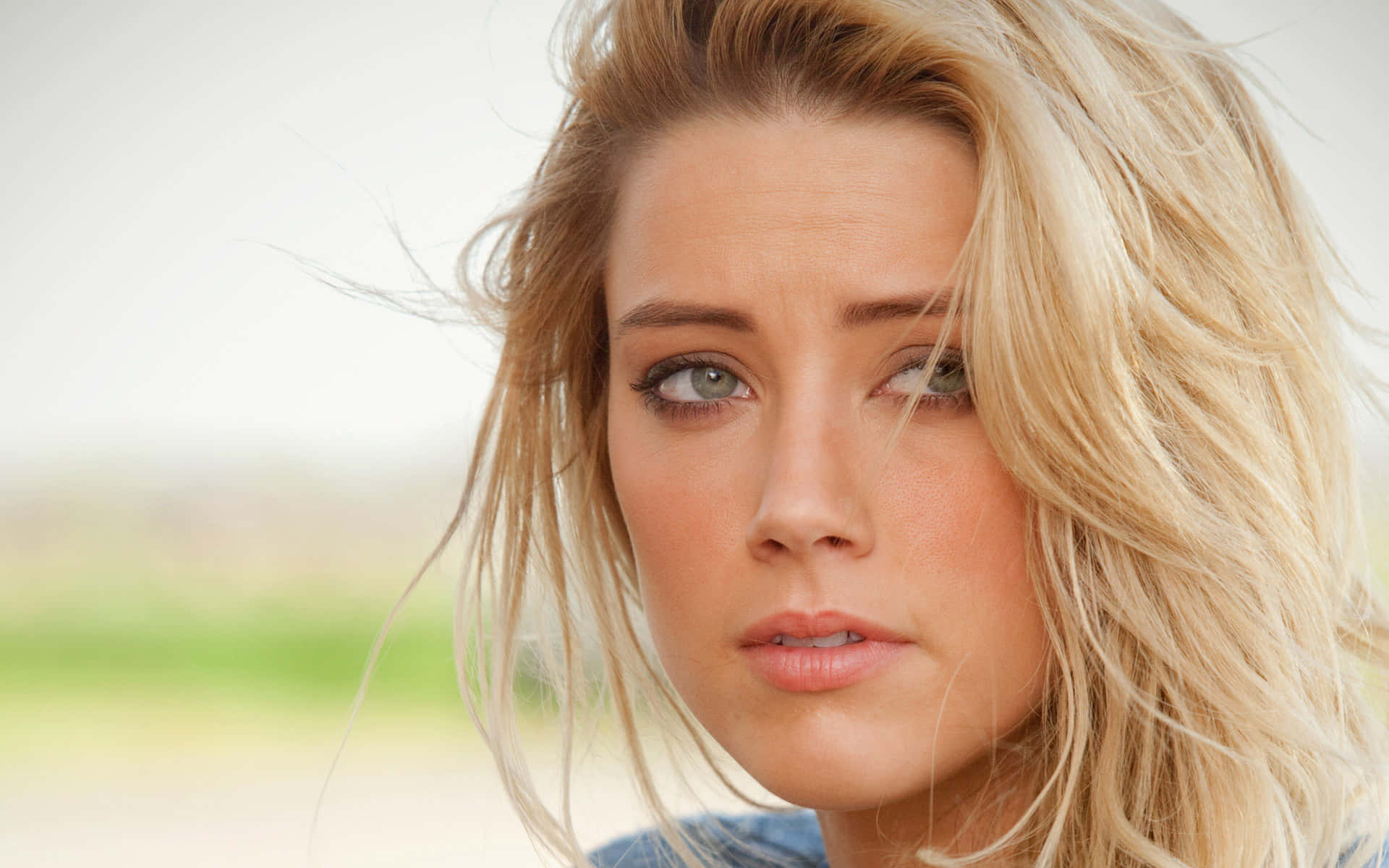 Amber Heard 桌布 壁紙 — 一位金髮女子的特寫肖像，頭髮被風吹起，在柔和模糊的戶外背景下若有所思地凝視。