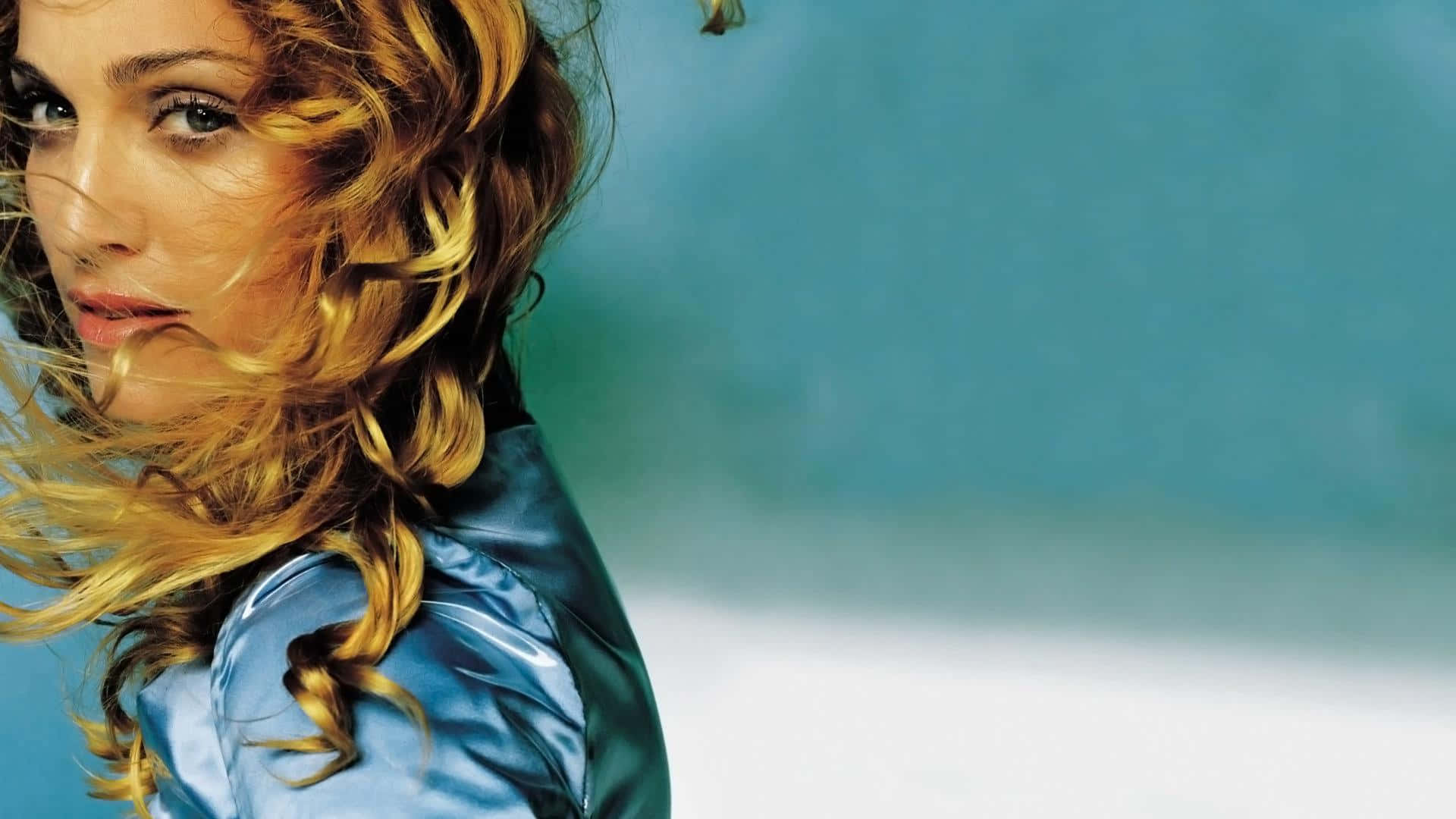 Madonna 桌布 壁紙 — 一位金髮女子眼神銳利，身穿閃亮藍色外套，背景為柔和青綠色和白色。