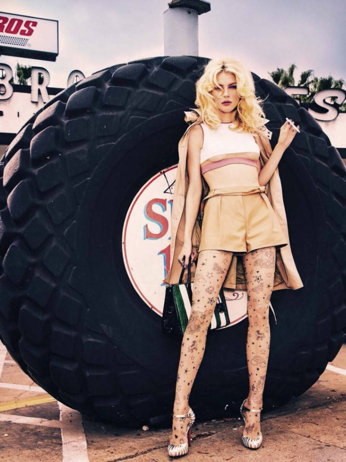 jessica stam 桌布 壁紙 — 一位金髮女子穿著復古風格服裝，在加油站自信地站在巨型怪獸卡車輪胎旁擺姿勢。