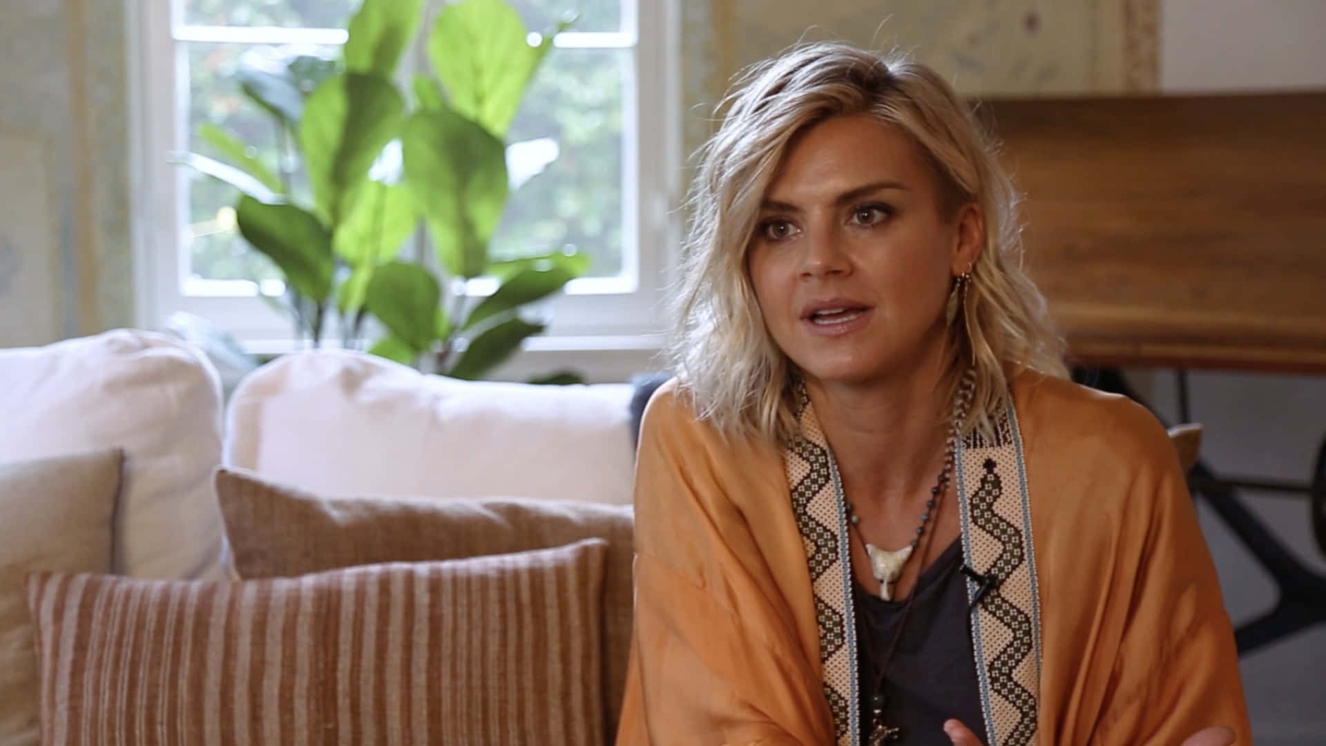 eliza coupe 桌布 壁紙 — 一位金髮女子穿著橙色和服，坐在有條紋靠墊的沙發上，身後有一棵大型綠色植物。