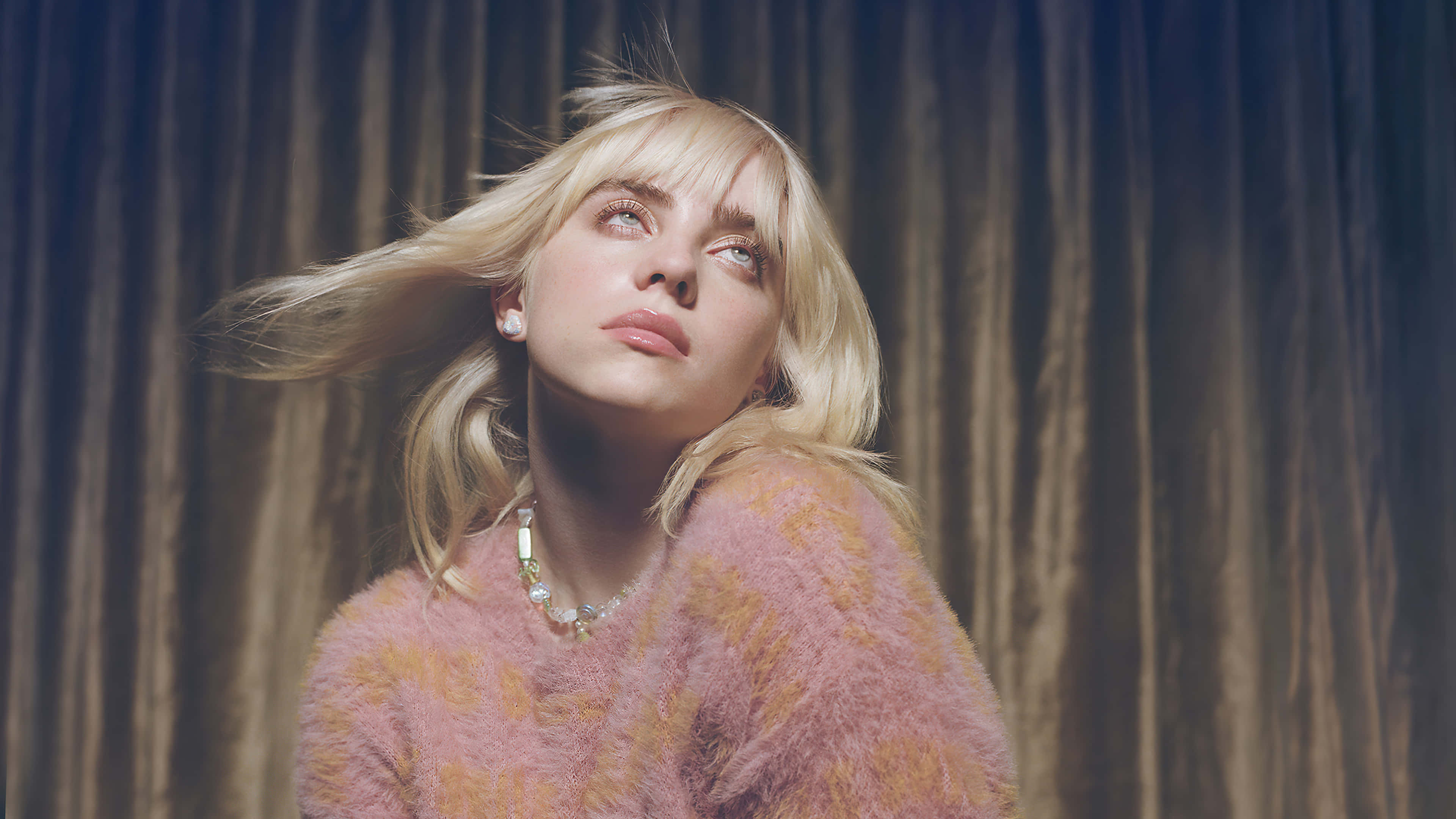 Billie Eilish 2021 桌布 壁紙 — 一位金髮女子穿著毛茸茸的粉紅色毛衣，在垂簾背景下，頭髮被風吹起，向上凝視。