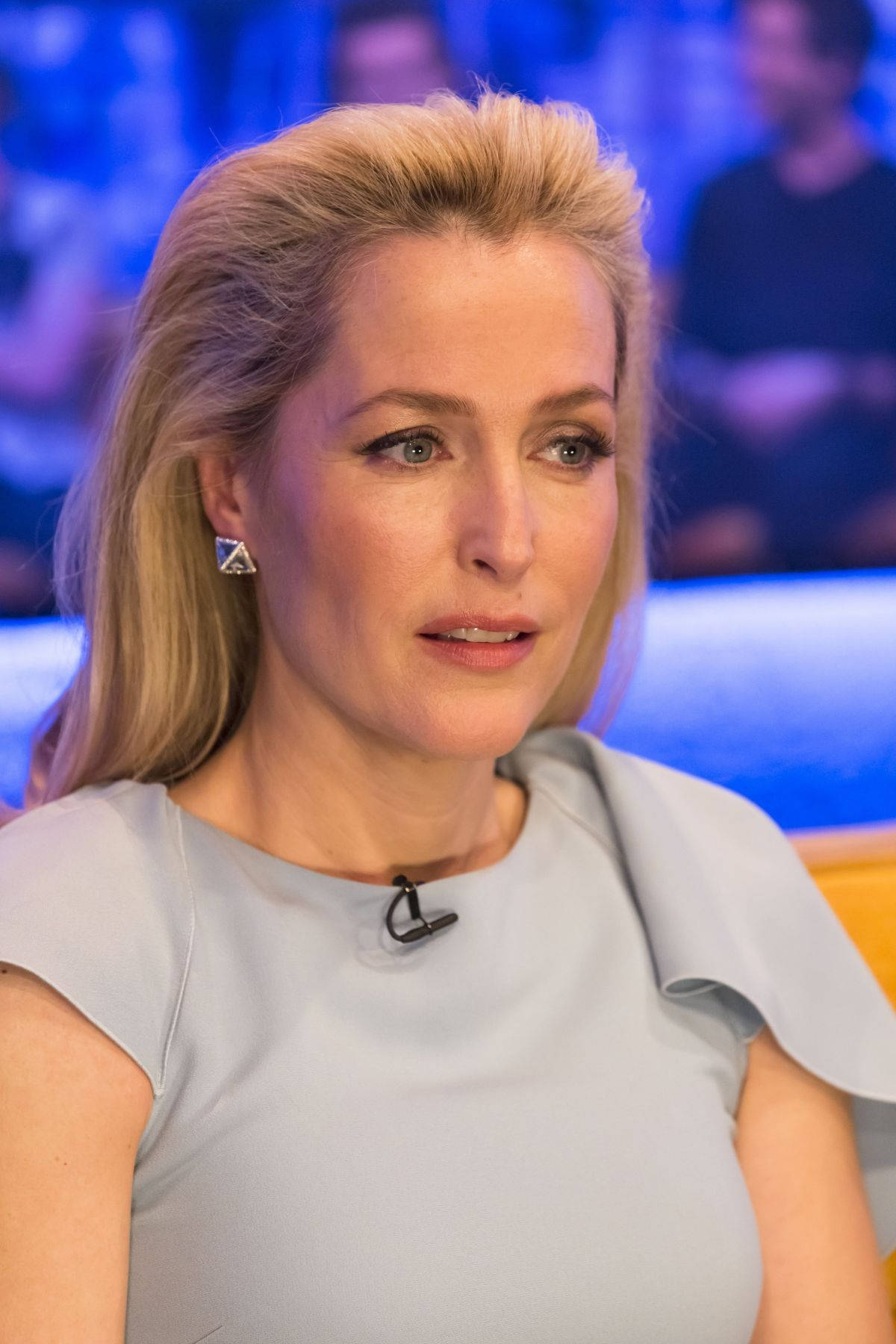 gillian anderson 桌布 壁紙 — 一位金髮女子穿著淺灰色連衣裙，坐在藍色燈光、背景有模糊觀眾的攝影棚中。