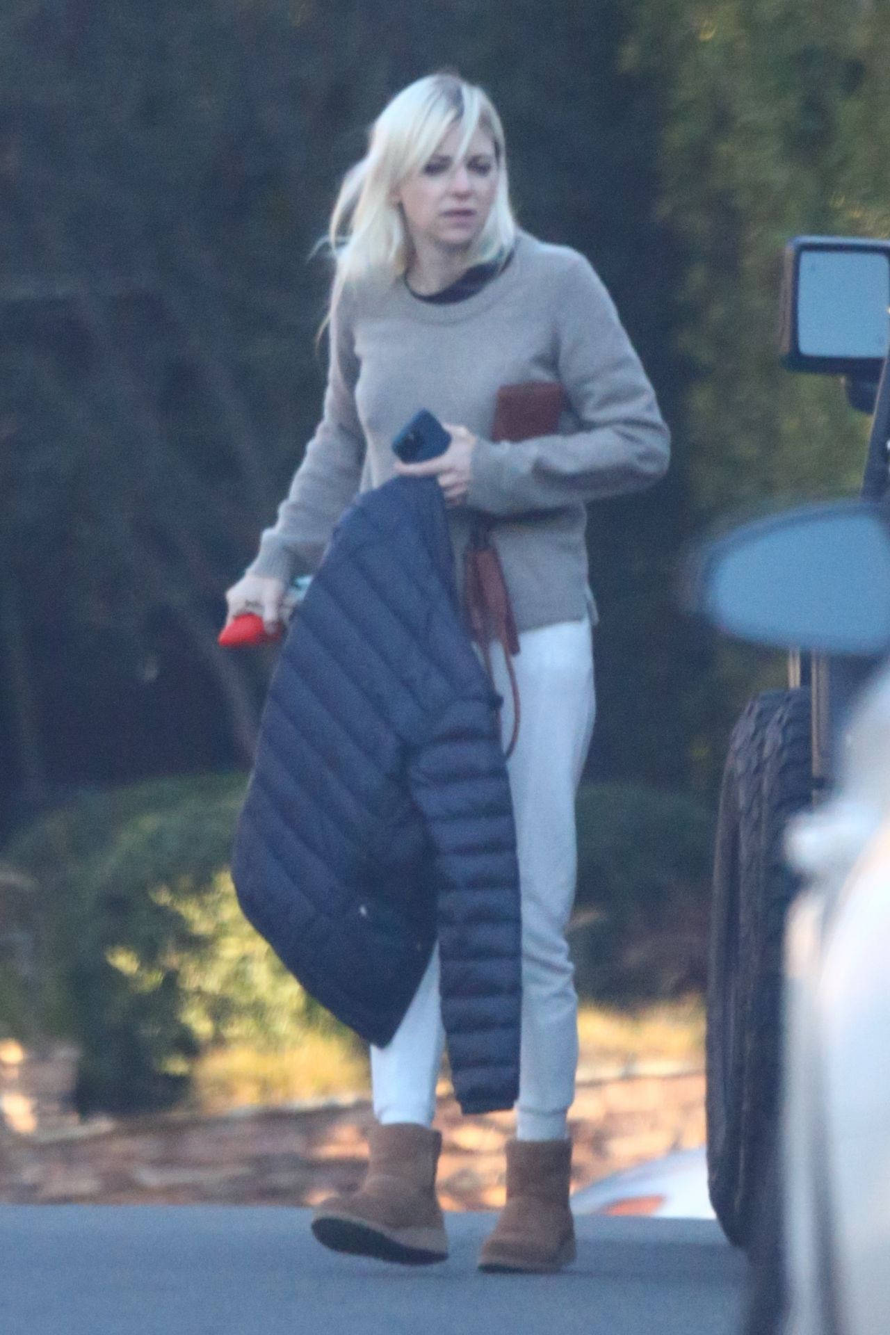 Celebrity 桌布 壁紙 — 一位金髮女子穿著灰色毛衣和白色長褲，在戶外行走，手持一件深色羽絨外套和一部手機。