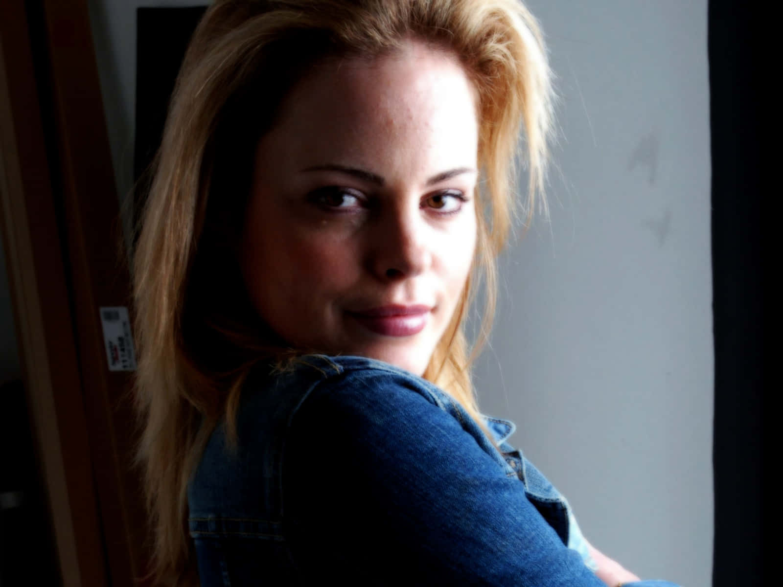 Chandra West 桌布 壁紙 — 一位金髮女子穿著牛仔外套，在昏暗的房間裡微笑著回頭看。