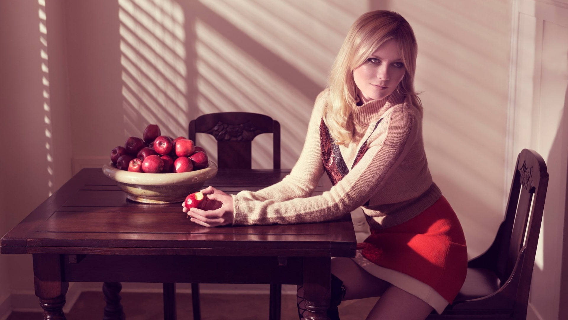 Kirsten Dunst 桌布 壁紙 — 一位金髮女子穿著米色毛衣和紅色裙子，坐在木桌旁，手裡拿著一個蘋果，旁邊有一碗蘋果，在溫暖的窗光下。