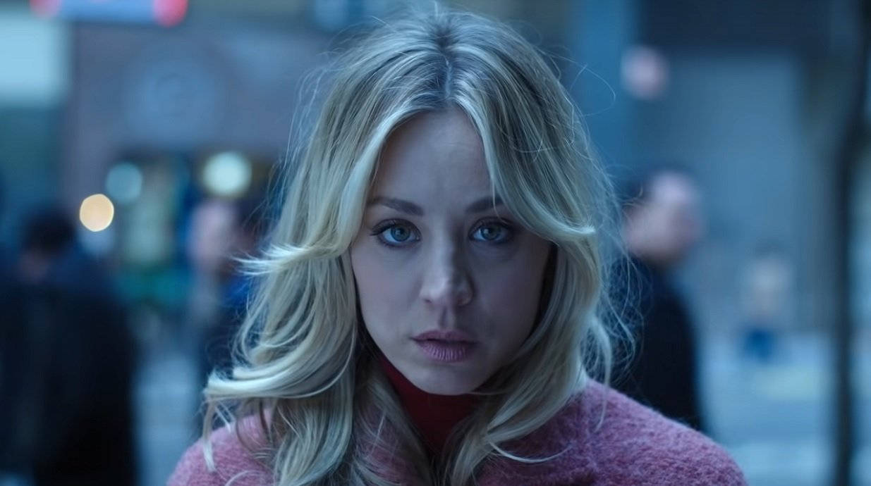 Kaley Cuoco 桌布 壁紙 — 一位金髮女子穿著粉紅色毛衣，在夜晚模糊的城市街道上專注地凝視著鏡頭。
