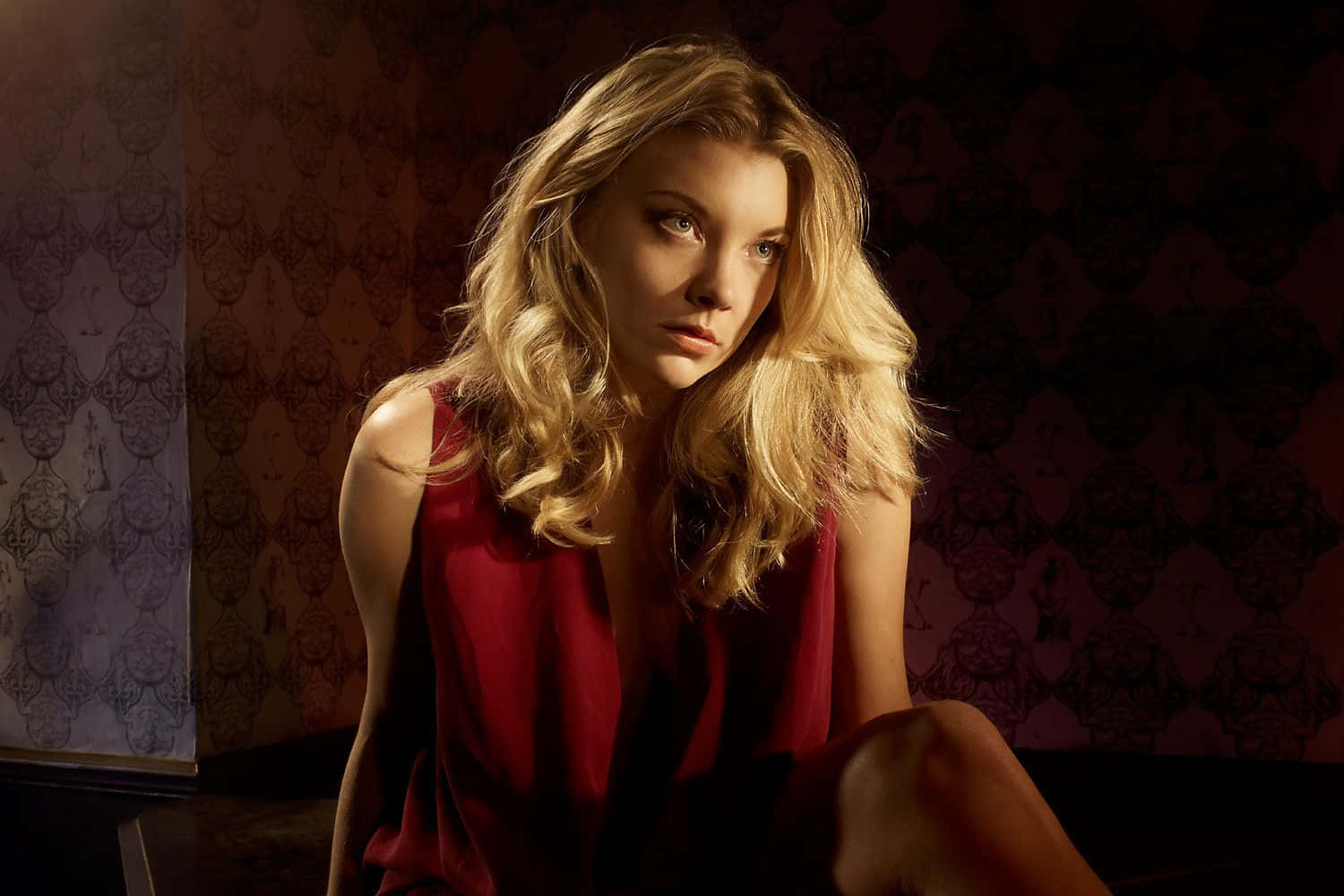 natalie dormer 桌布 壁紙 — 一位金髮女子穿著紅色連衣裙，在昏暗光線下坐在華麗壁紙前，神情嚴肅。