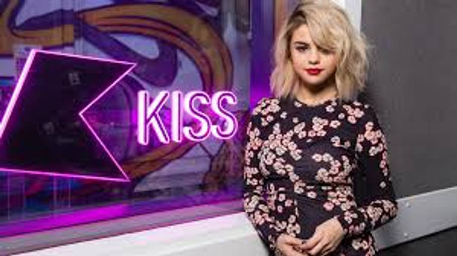 Selena Gomez 桌布 壁紙 — 一位金髮女子穿著花卉連衣裙，站在現代工作室環境中發光的粉紅色霓虹燈 'KISS' 標誌前。