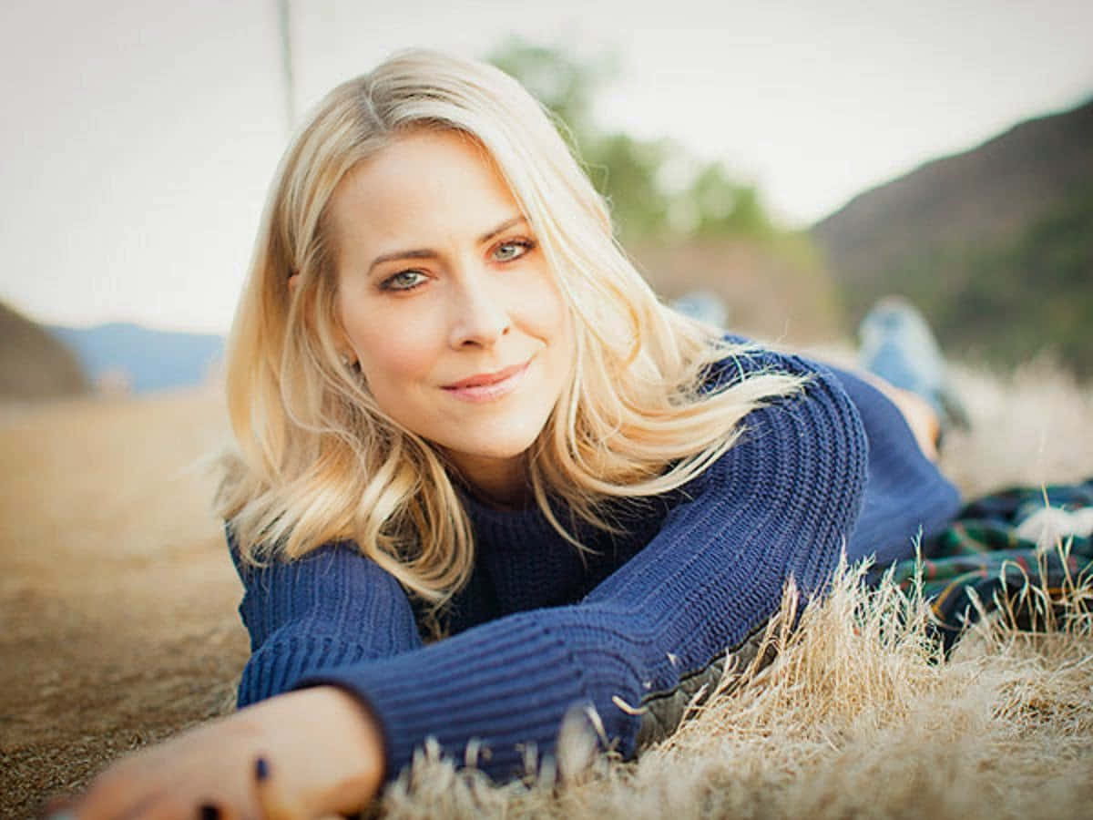 Brittany Daniel 桌布 壁紙 — 一位金髮女子穿著藍色毛衣，躺在乾草地上，背景有山丘，露出溫柔的微笑。
