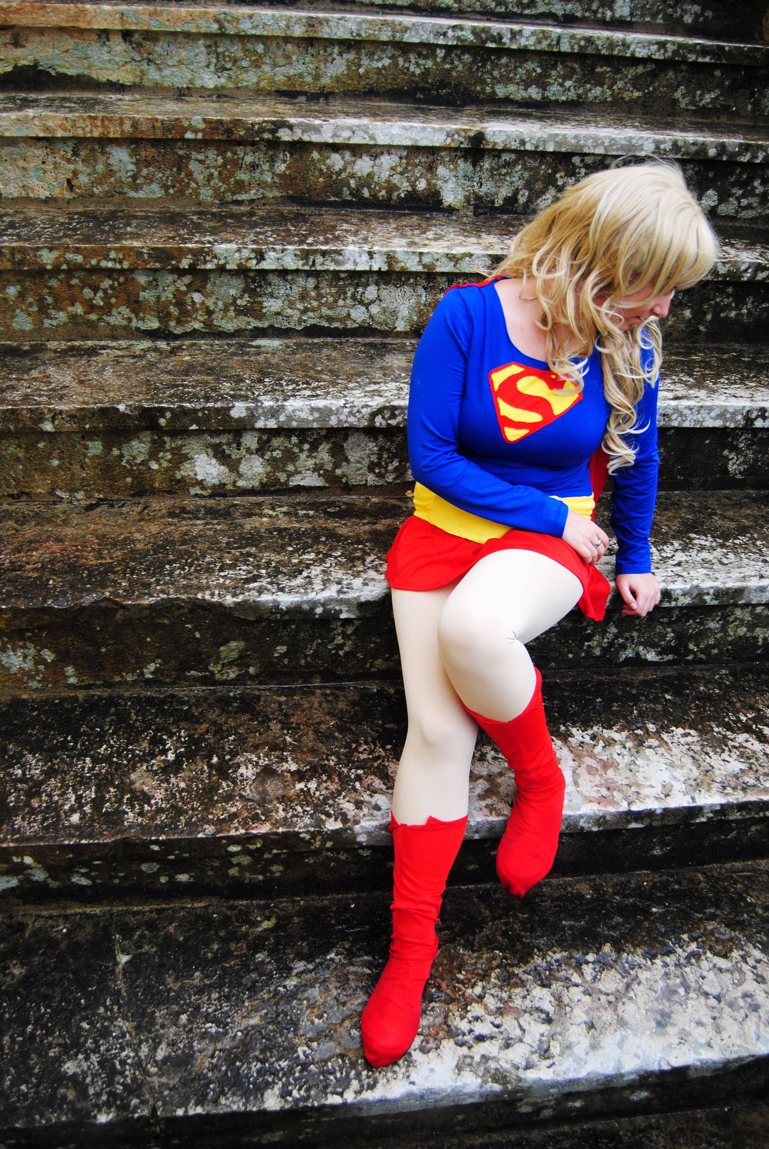 Superwoman 桌布 壁紙 — 一位金髮女子穿著超人服裝，低頭沉思地坐在風化的石階上，周圍佈滿苔蘚和鐵鏽。