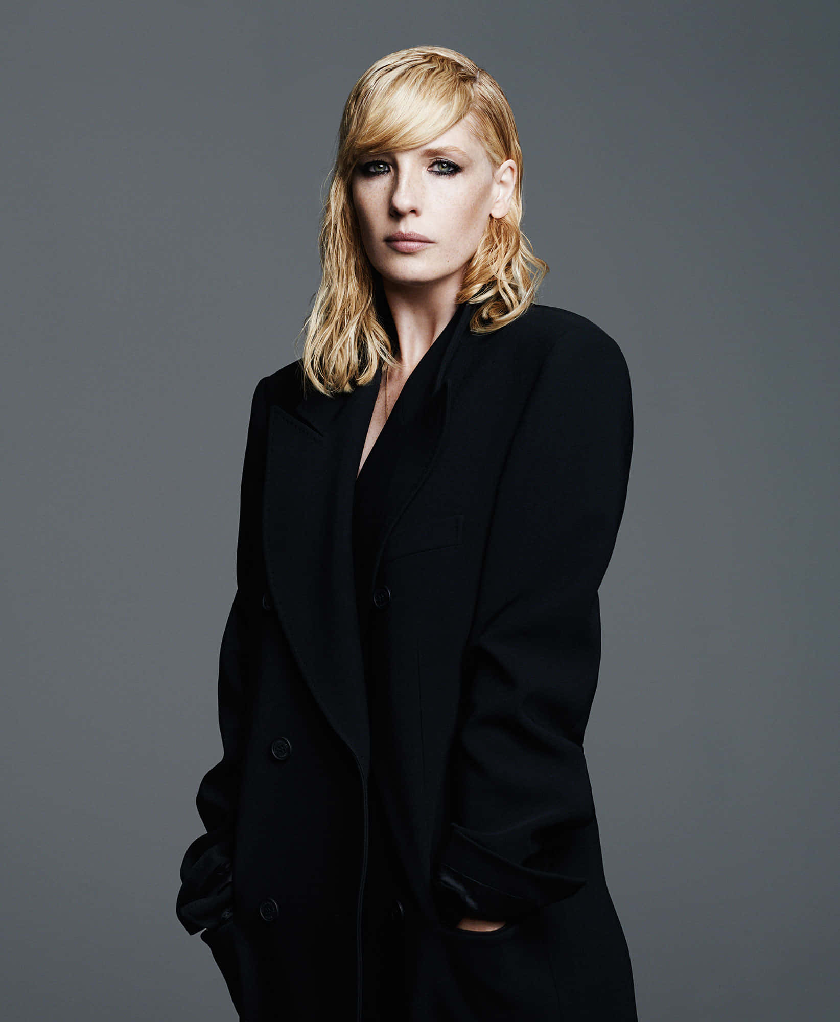 Kelly Reilly 桌布 壁紙 — 一位金髮女子穿著黑色外套，在灰色背景前表情嚴肅地站立。