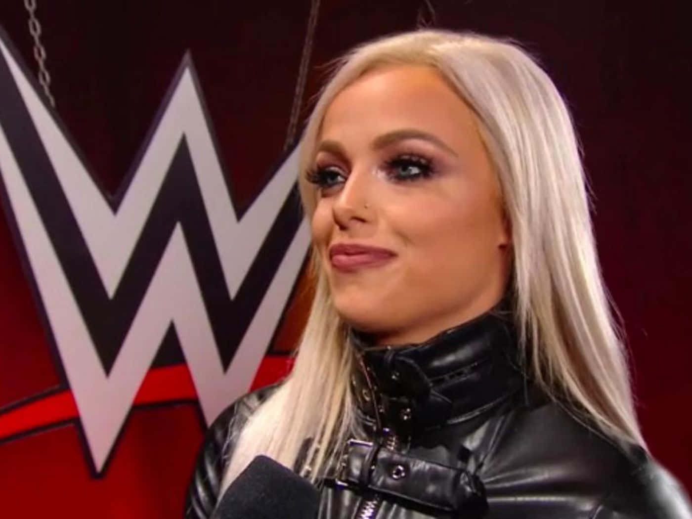 WWE 桌布 壁紙 — 一位金髮女子穿著黑色皮夾克，在WWE標誌前對著麥克風微笑說話。