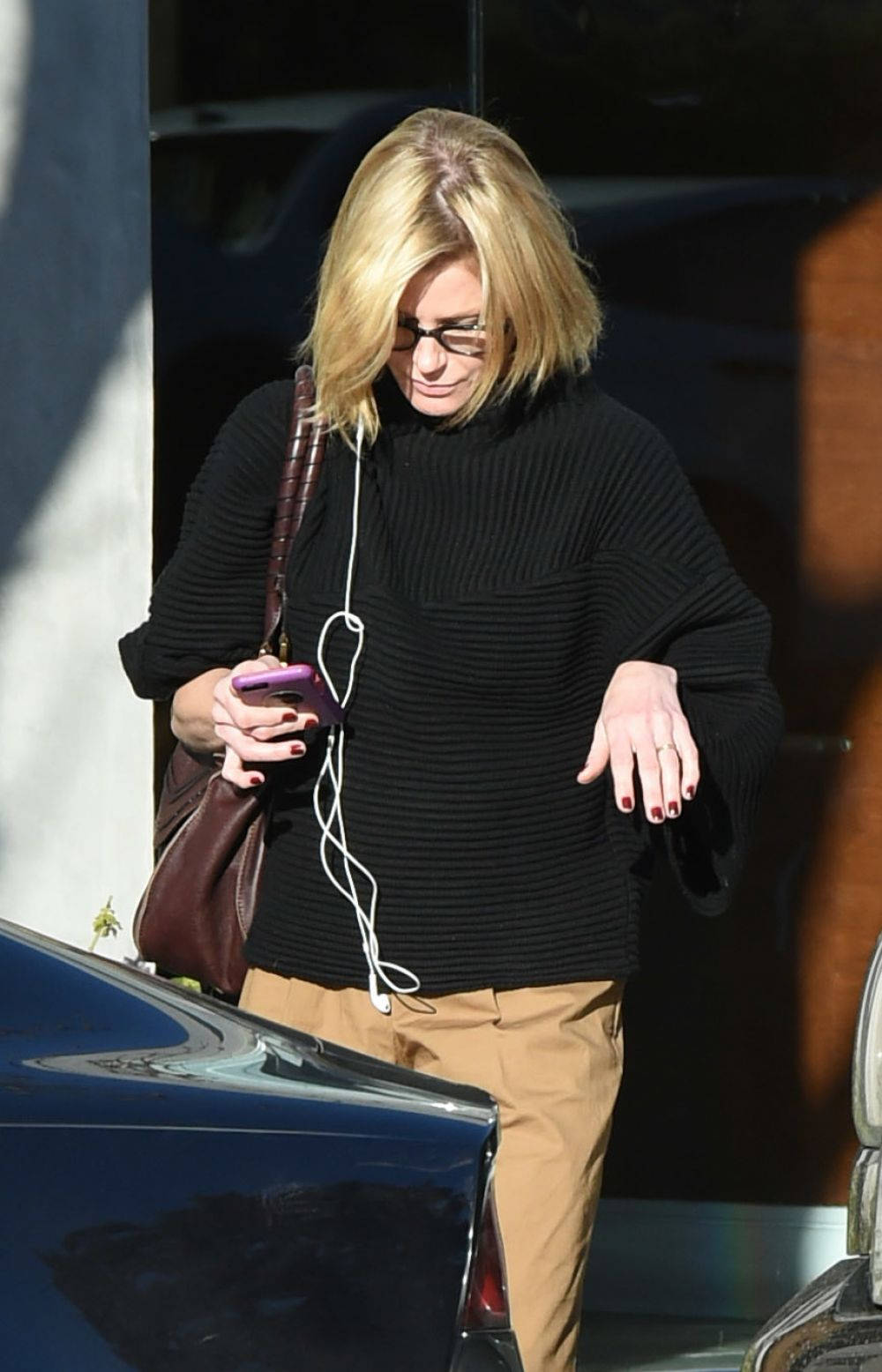 Celebrity 桌布 壁紙 — 一位金髮女子穿著黑色羅紋毛衣和眼鏡，在戶外行走時看著她的紫色手機，白色耳機垂掛著。