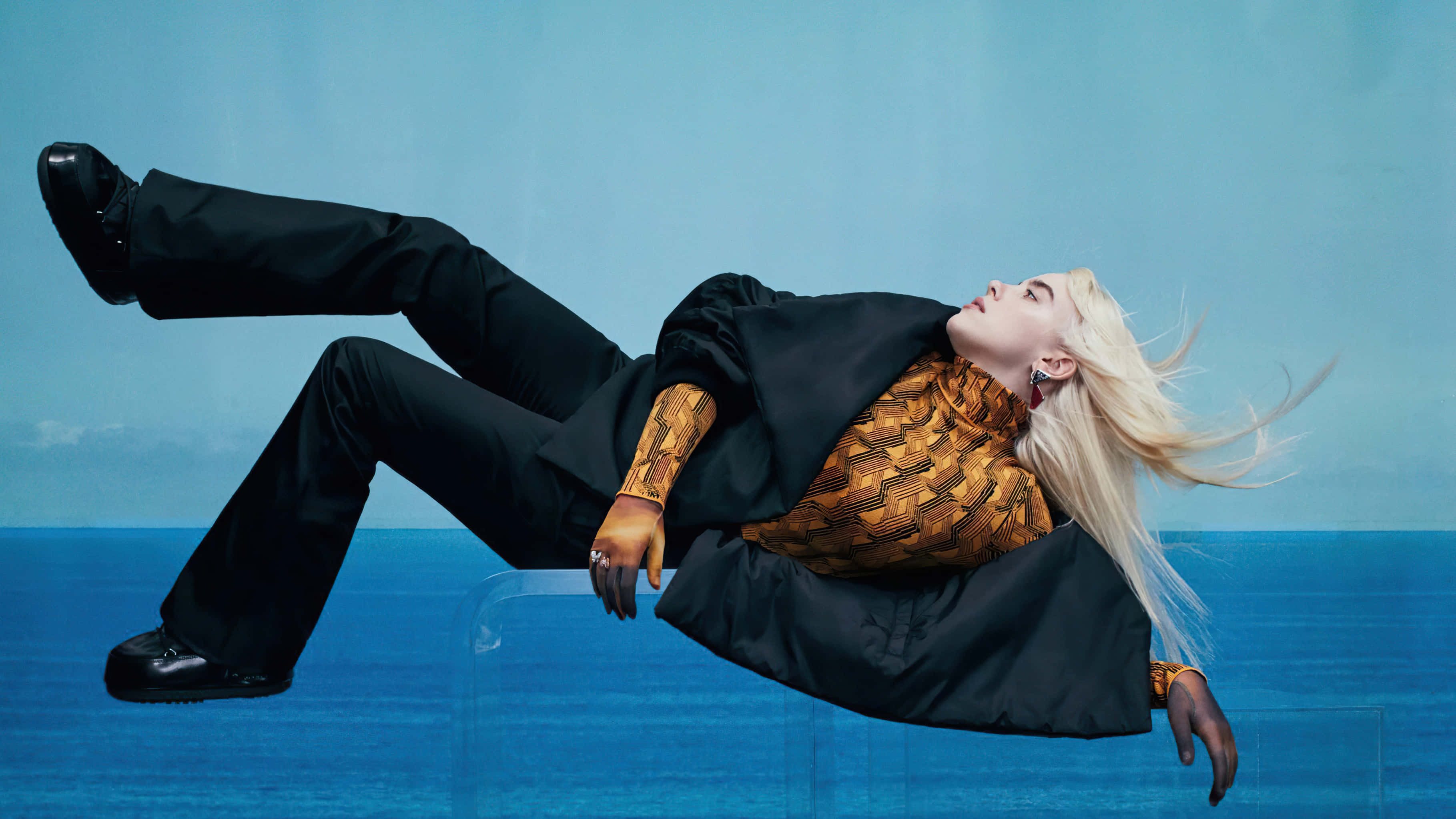 Billie Eilish 2021 桌布 壁紙 — 一位金髮女子穿著黑色褲子和圖案上衣，在藍色漸變背景和下方海洋的半空中漂浮。
