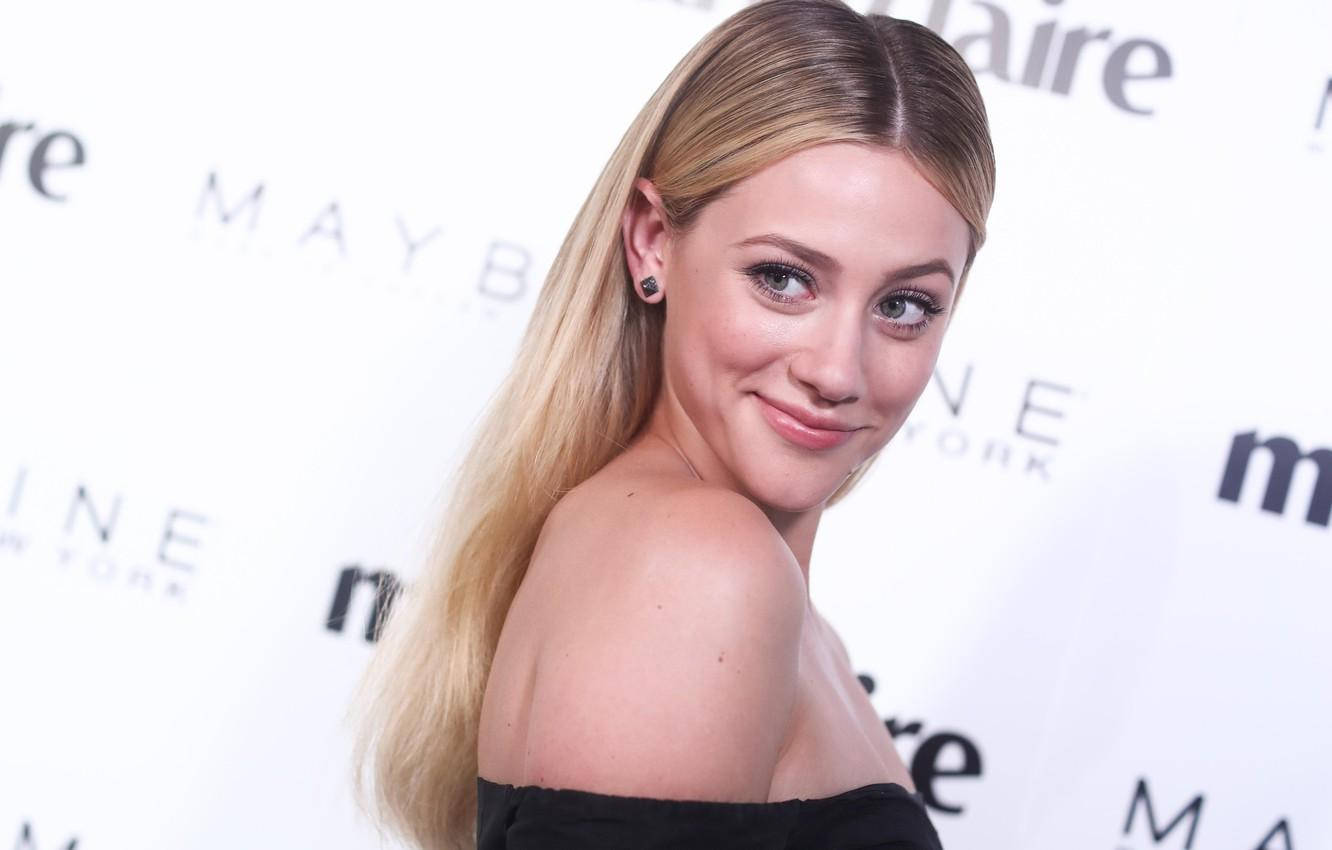 Celebrity 桌布 壁紙 — 一位金髮女子穿著黑色露肩連衣裙，越過肩膀對著白色品牌活動背景微笑。